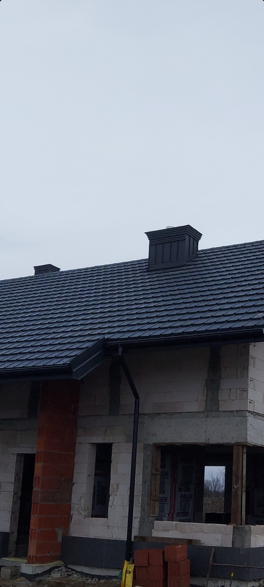 Nowy dach z ciemnej dachówki ceramicznej i czarna rynna na budynku w trakcie budowy, widoczne kominy.