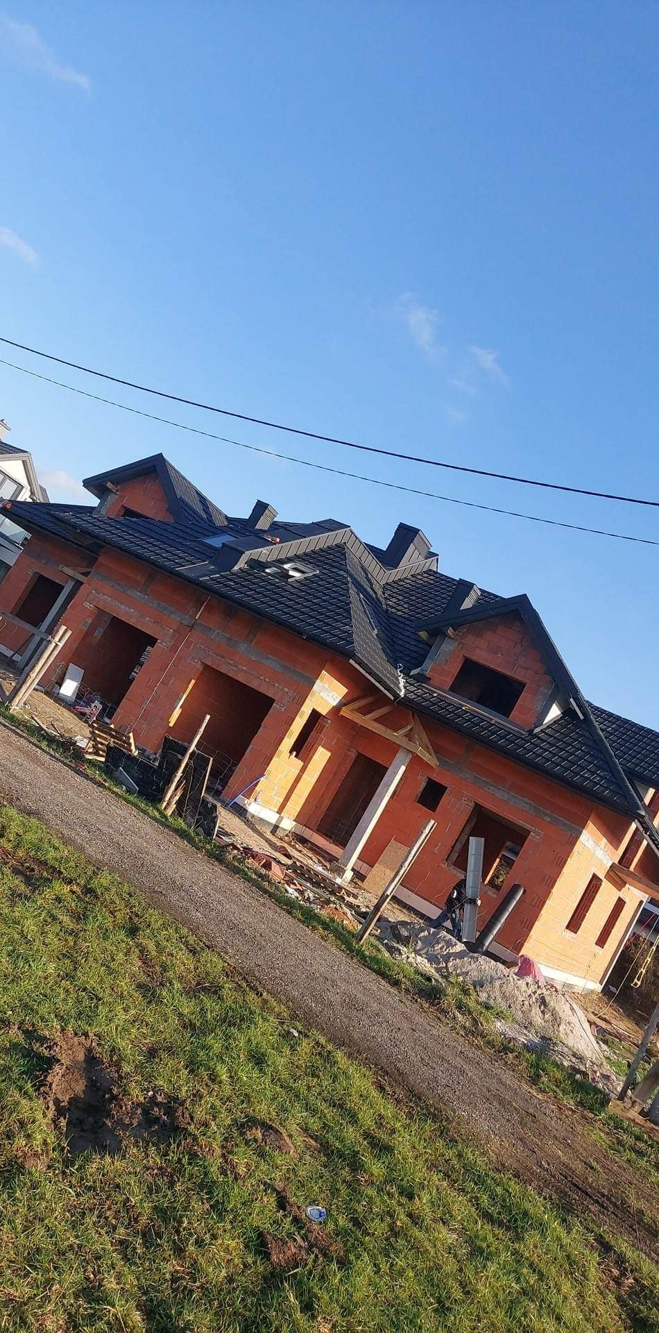 Nowo kryty dach z czarnej dachówki na budynku w stanie surowym otwartym, widok z poziomu gruntu na tle błękitnego nieba.