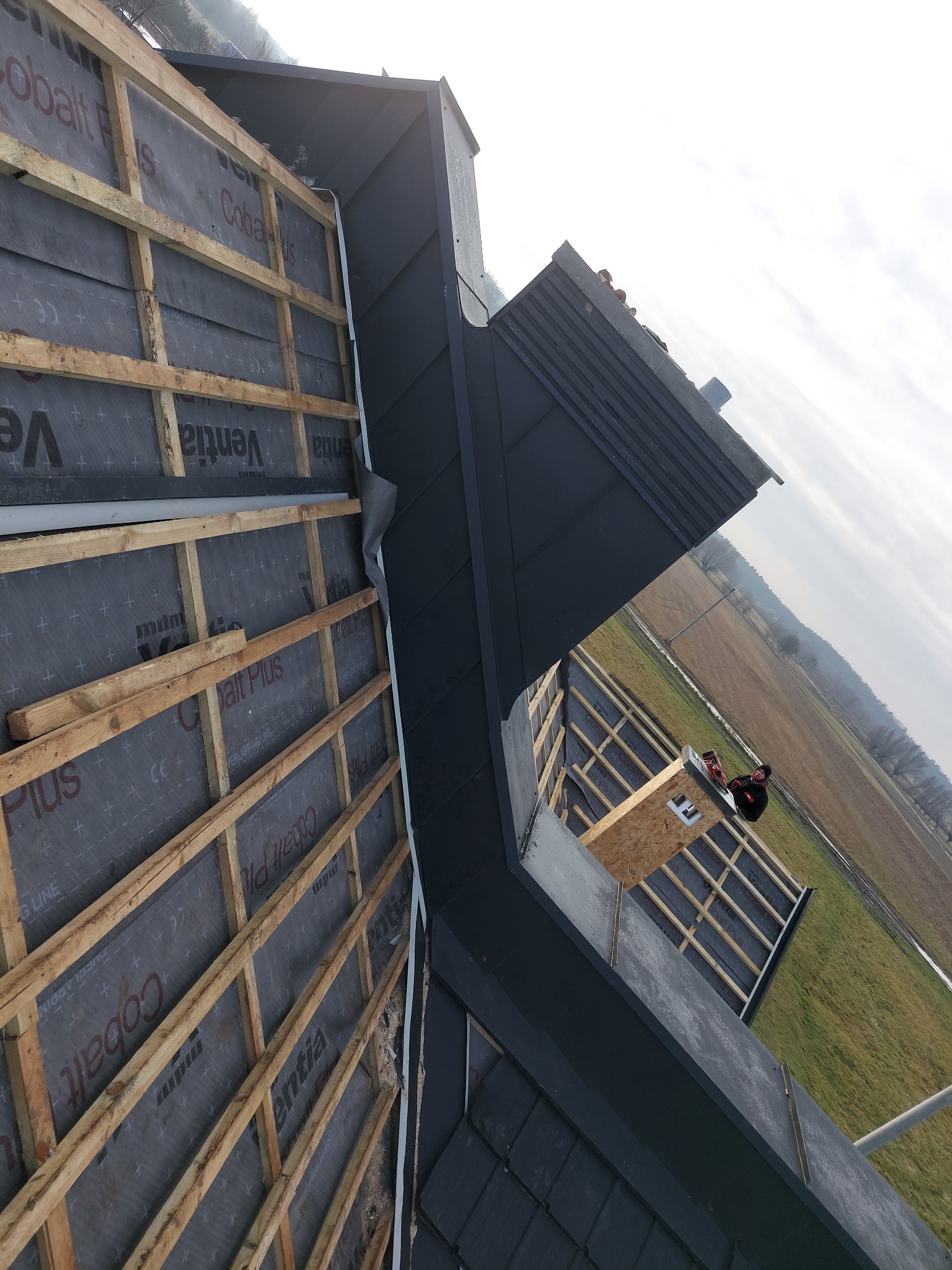 Częściowy widok dachu z drewnianą konstrukcją, membraną i blachą; w tle krajobraz z polami i pracownik na dachu dobudówki.