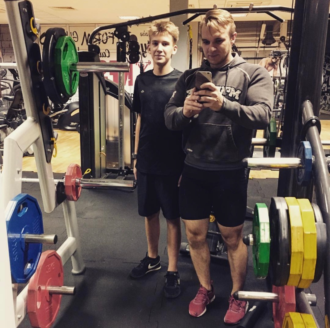 Mężczyzna w bluzie z kapturem robi selfie z nastolatkiem na siłowni w Kielcach, obok stojak na ciężary z kolorowymi obciążeniami.