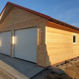 Drewniany garaż dwustanowiskowy z jasnymi, segmentowymi bramami, brązowym dachem i rynną, oświetlony popołudniowym słońcem.