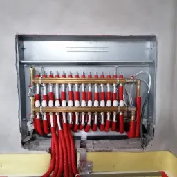 Rozdzielacz ogrzewania podłogowego z rurami w izolacji czerwonej, zamontowany w metalowej szafce w surowej ścianie, widoczne elementy instalacji na podłodze z folią grzewczą.