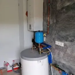 Biały piec gazowy z niebieskim zbiornikiem wyrównawczym i miedzianymi rurami zamontowany na szarej ścianie z płytek, widoczne elementy instalacji hydraulicznej i grzewczej oraz materiały budowlane...