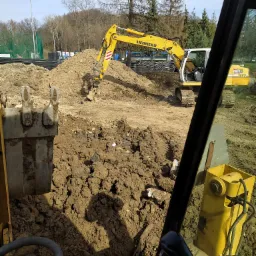 Widok z kabiny koparki na plac budowy z pracującą koparką Liebherr, sterta ziemi i fragmenty instalacji w tle.