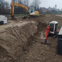 Dwa różne modele koparek (Liebherr i Kubota) pracują przy wykopie w ziemi, widoczna drabina oparta o skarpę, w tle zabudowania i drzewa w pochmurny dzień.