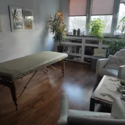 Jasny gabinet z kozetką do masażu, fotelami i stolikiem z owocami oraz bukietem tulipanów, na parapecie rośliny doniczkowe i książki.