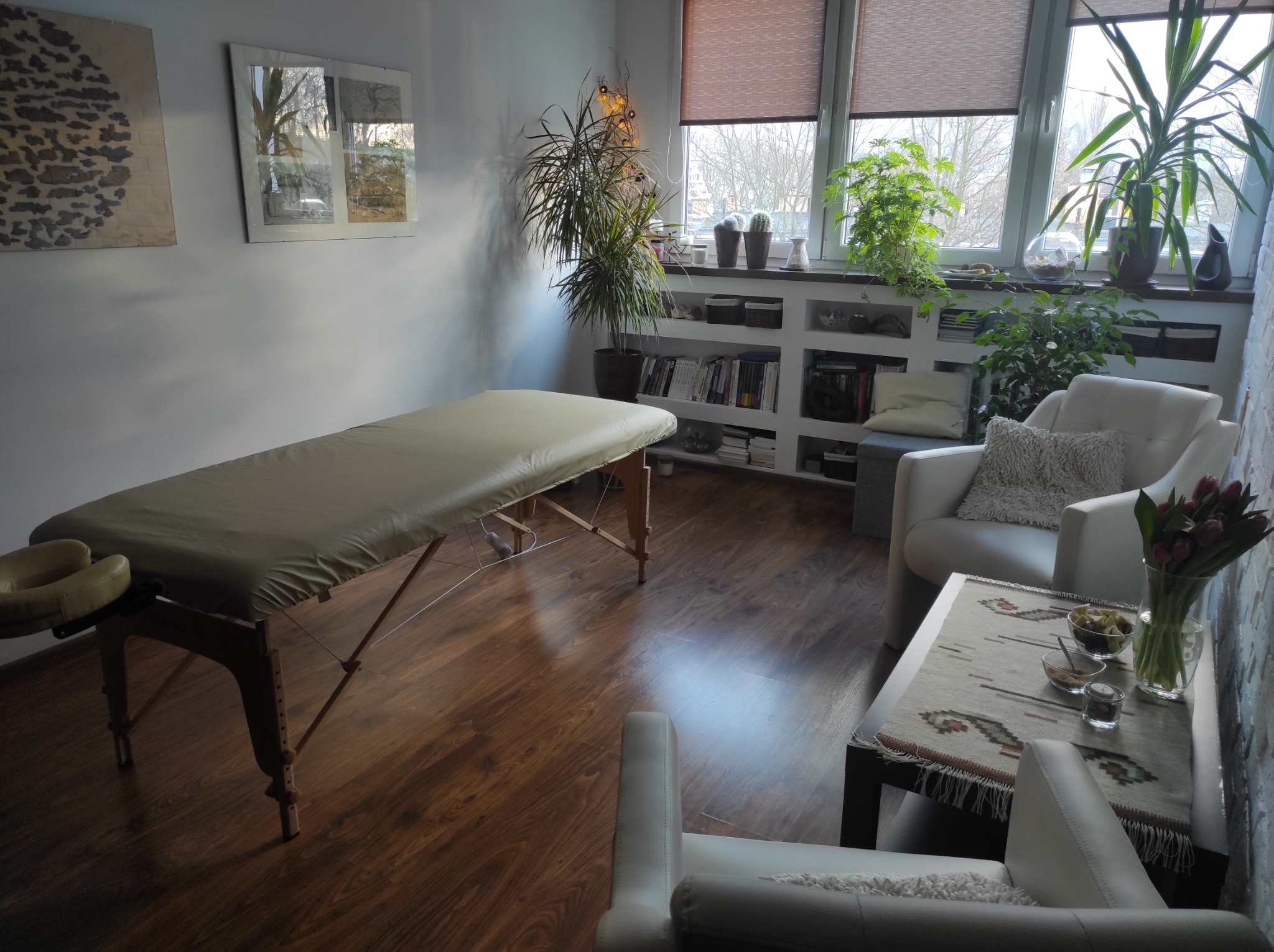 Jasny gabinet z kozetką do masażu, fotelami i stolikiem z owocami oraz bukietem tulipanów, na parapecie rośliny doniczkowe i książki.