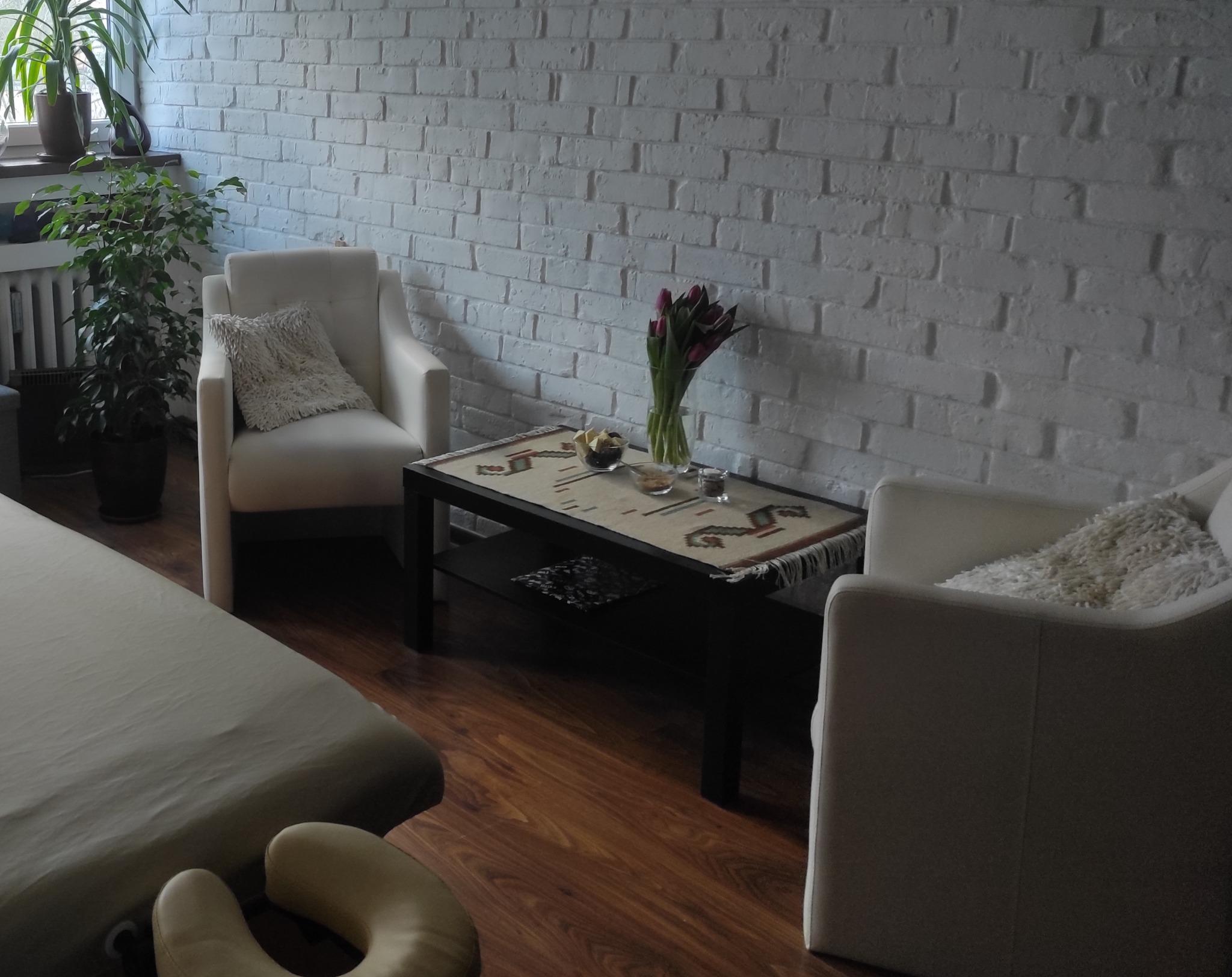 Jasne wnętrze gabinetu z kozetką, dwoma fotelami, stolikiem z kwiatami i przekąskami, oraz ścianą z białej cegły.