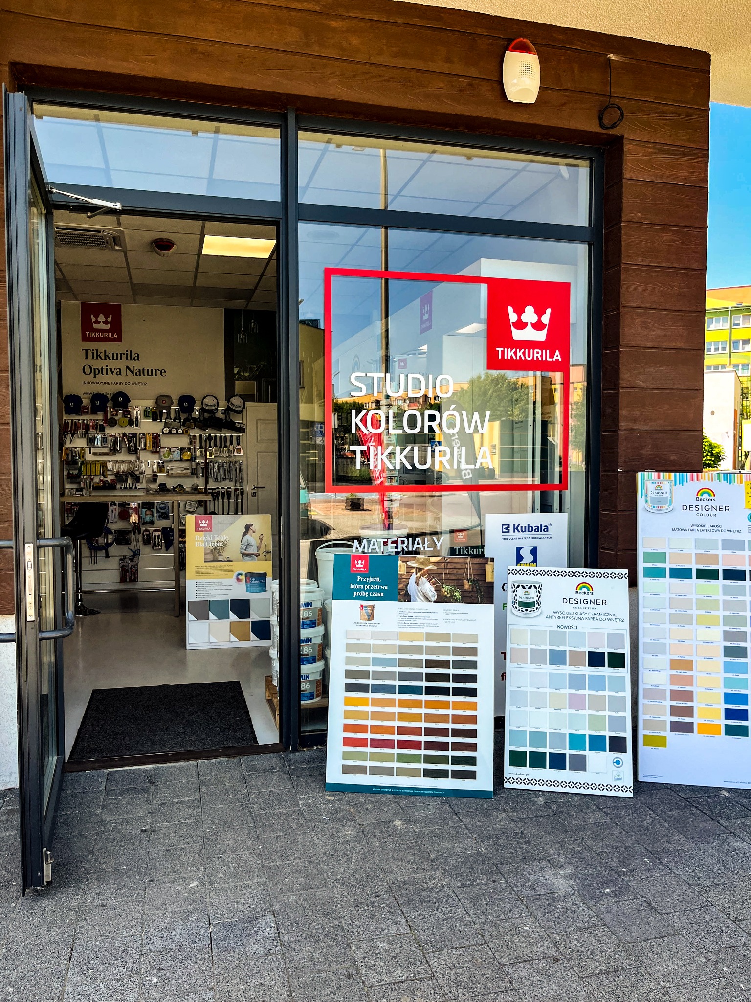 Wejście do sklepu z farbami Tikkurila, widoczne ekspozytory z paletami kolorów i narzędzia malarskie wewnątrz.