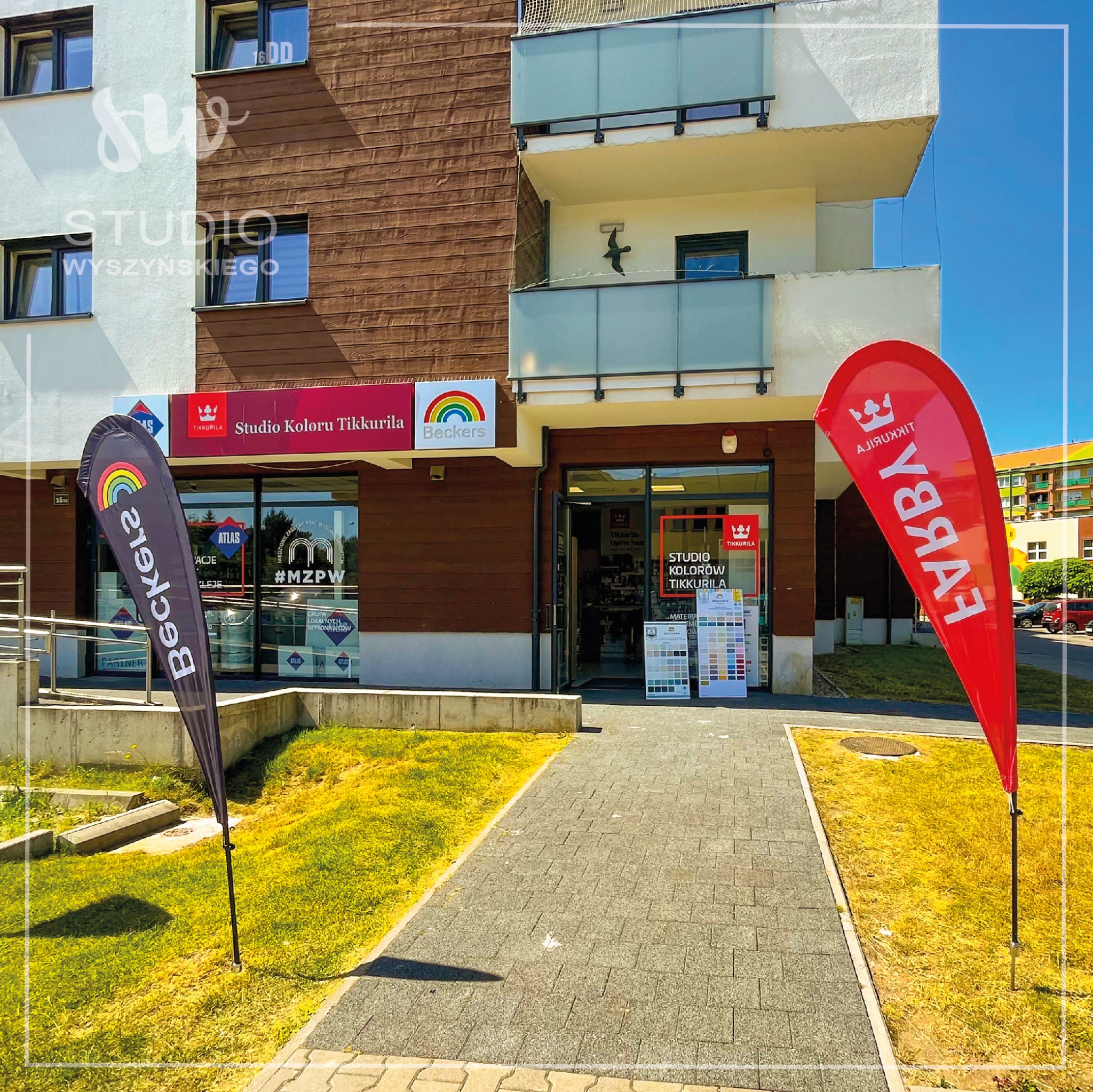 Fasada budynku z brązową drewnianą elewacją, przed wejściem stoją dwie flagi reklamowe marek Beckers i Tikkurila, widoczne okna z ekspozycją produktów i palet kolorów.