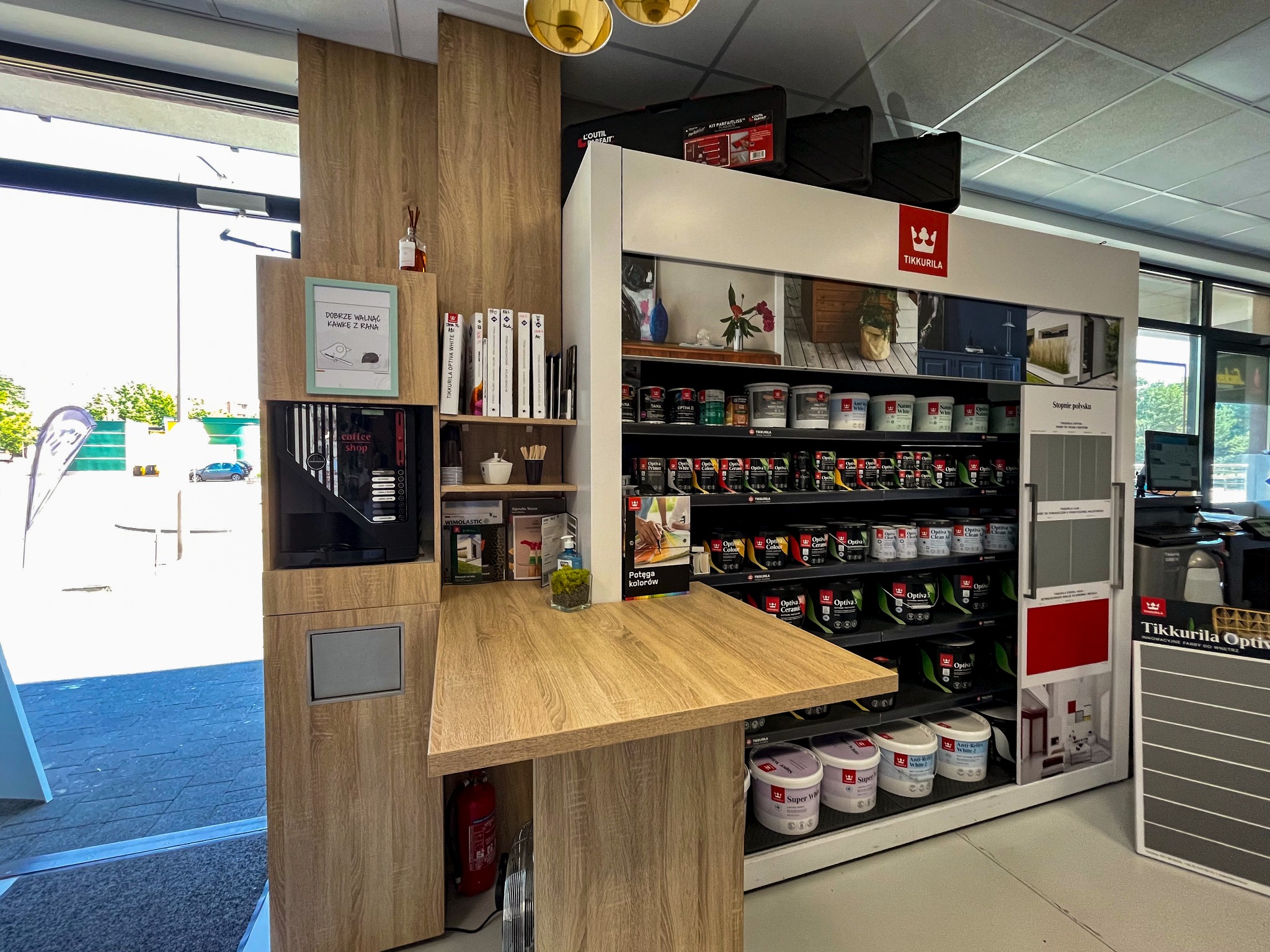Wnętrze sklepu budowlanego Tikkurila z ekspozycją farb, regałem z akcesoriami i kącikiem kawowym z plakatem 'Dobrze walnąć kawę z rana'.