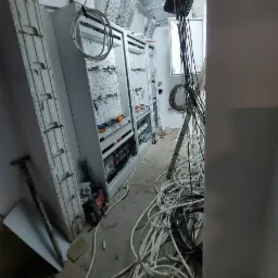 Dwa otwarte panele elektryczne w trakcie montażu w surowym pomieszczeniu, widoczne kable, drabina i narzędzia.