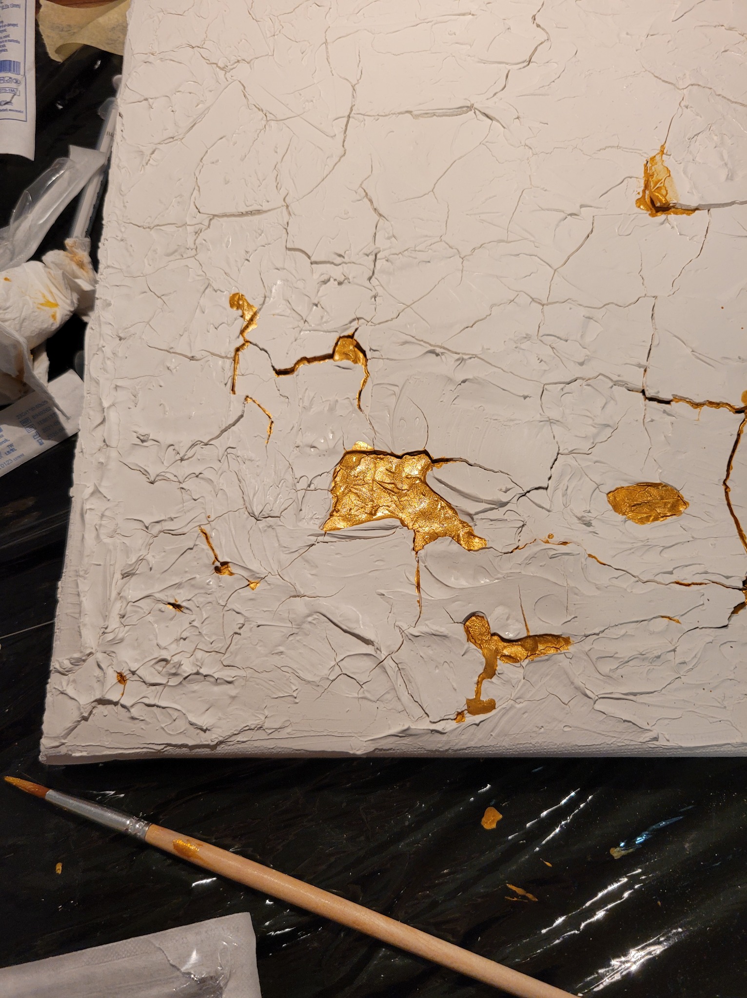 Biała, teksturowana powierzchnia z nieregularnymi pęknięciami wypełnionymi złotą farbą, widoczny pędzel z resztkami złotej farby na czarnym tle.