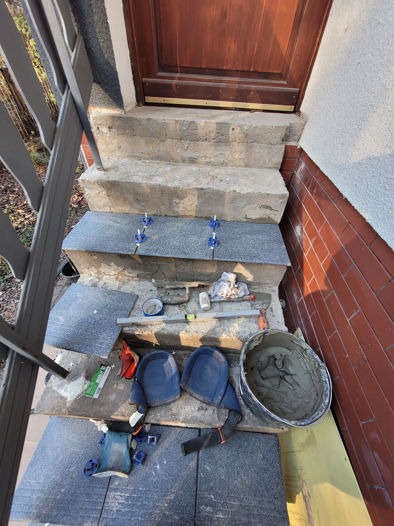 Montaż płytek na schodach wejściowych, widoczne narzędzia glazurnicze, zaprawa w wiadrze, poziomica oraz płytki z niebieskimi krzyżykami dystansowymi.