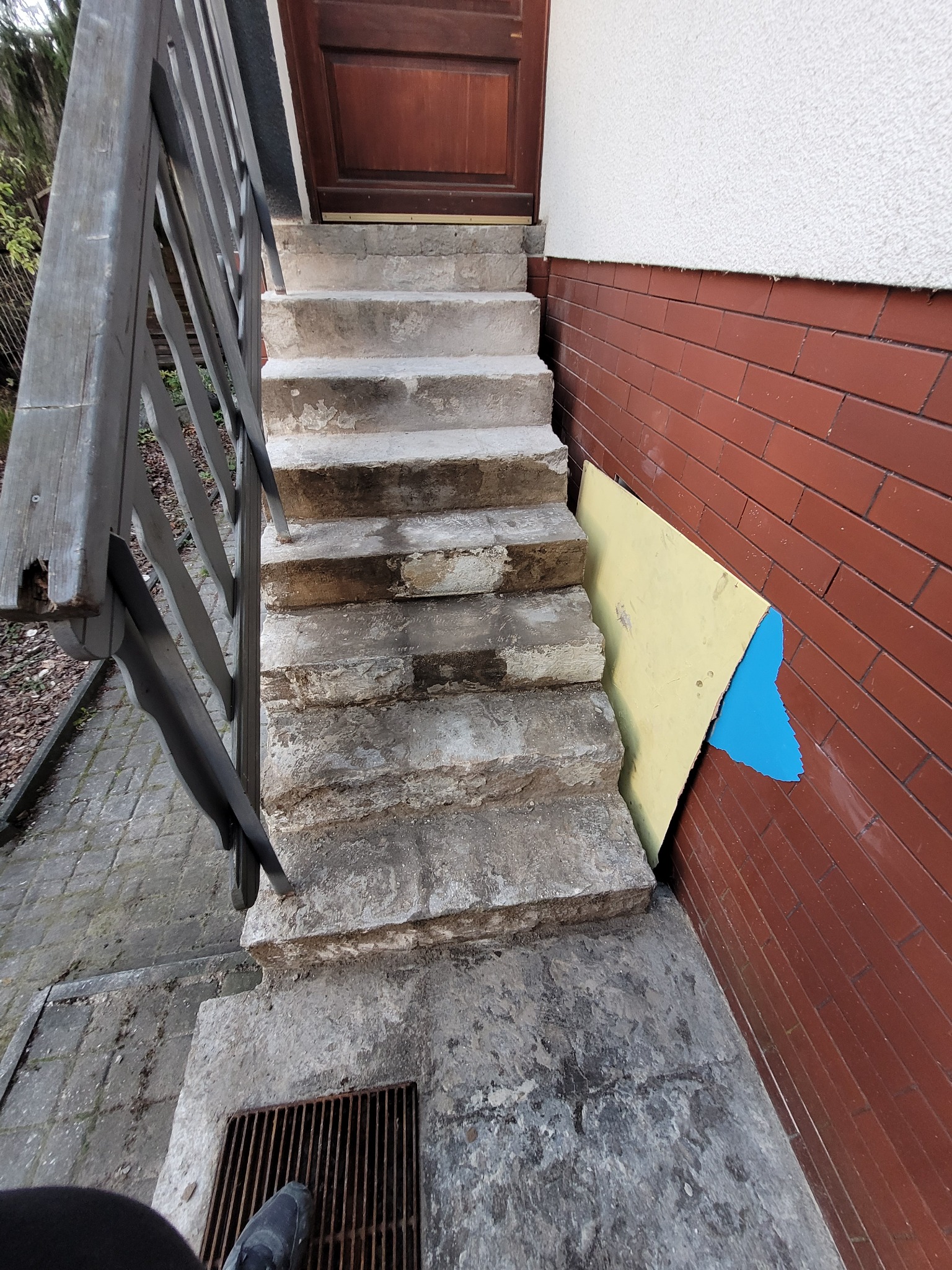 Zewnętrzne, betonowe schody wejściowe do domu, z widocznymi śladami uszkodzeń i prowizorycznym zabezpieczeniem ściany płytą z resztkami farby. Schody prowadzą do brązowych drzwi, a obok znajduje...