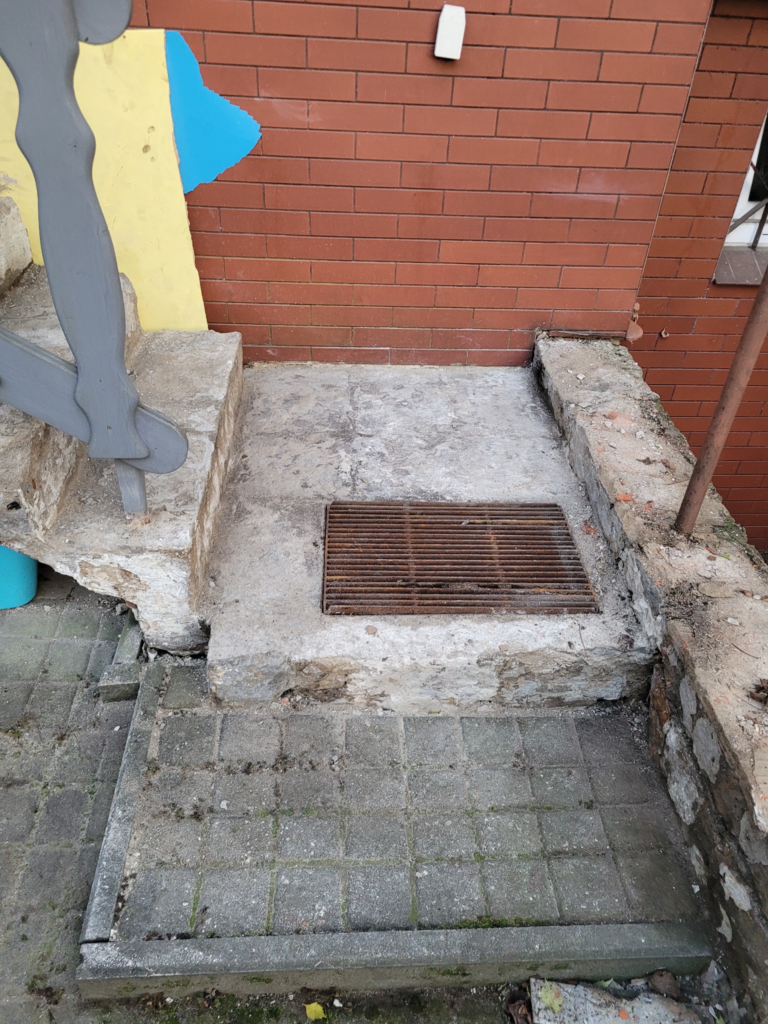 Zniszczone betonowe schody z metalową kratką odpływową w Poznaniu, przylegające do ceglanej ściany z fragmentem żółto-niebieskiej dekoracji.