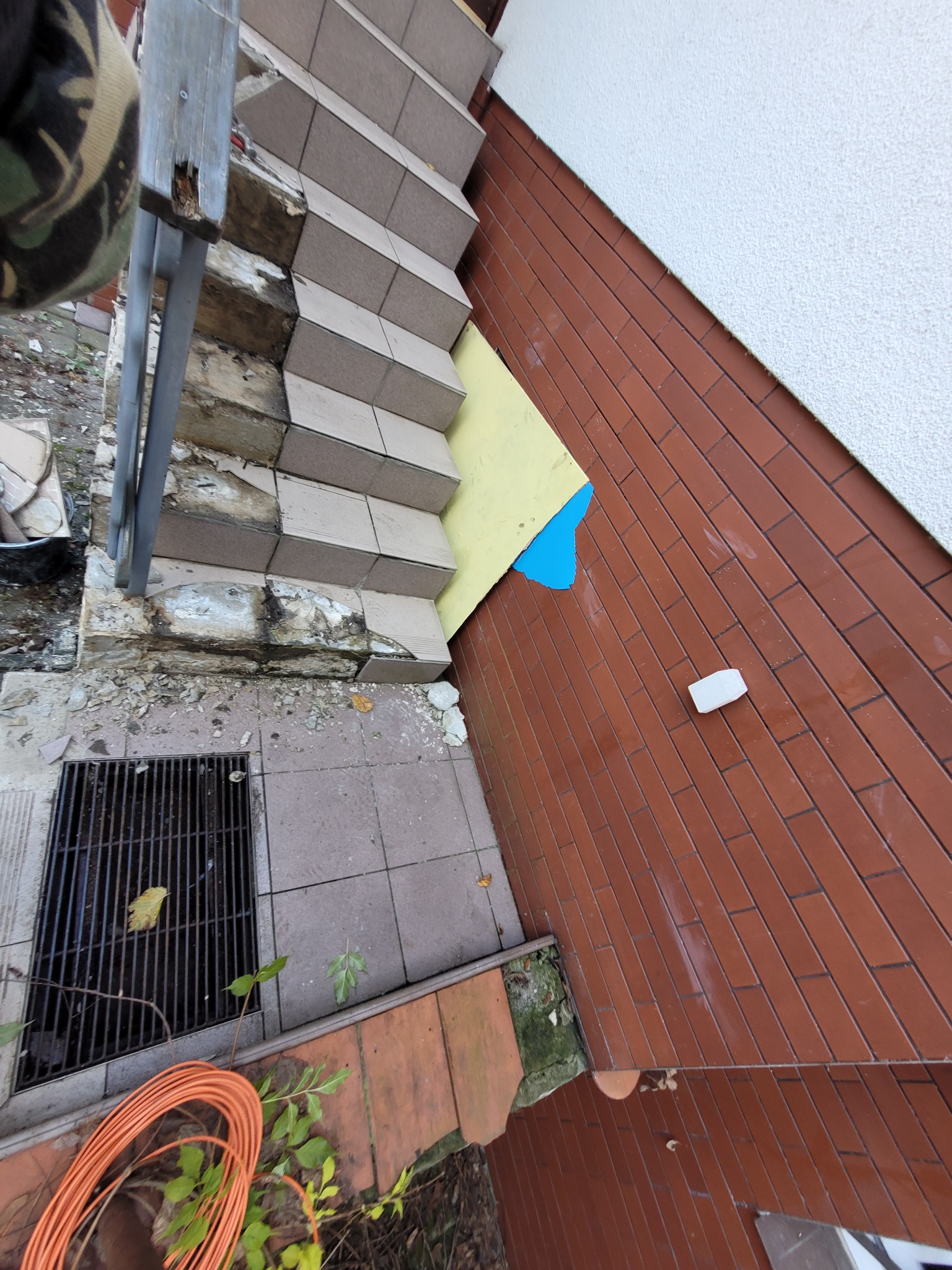Zniszczone schody zewnętrzne z odpadającym tynkiem i płytkami, widoczne przygotowania do naprawy z żółtą i niebieską folią ochronną na ścianie.