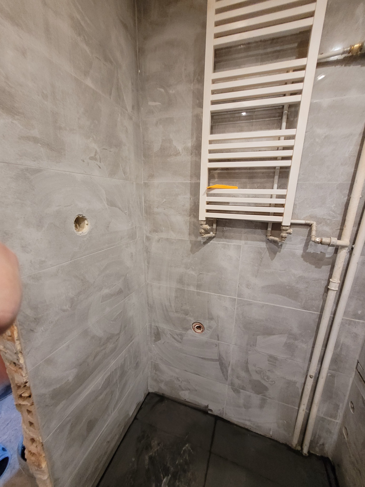 Wnętrze łazienki w trakcie remontu: szare płytki imitujące beton, zamontowany biały grzejnik drabinkowy z żółtą poziomnicą, widoczne rury i otwory w ścianie przed montażem armatury.