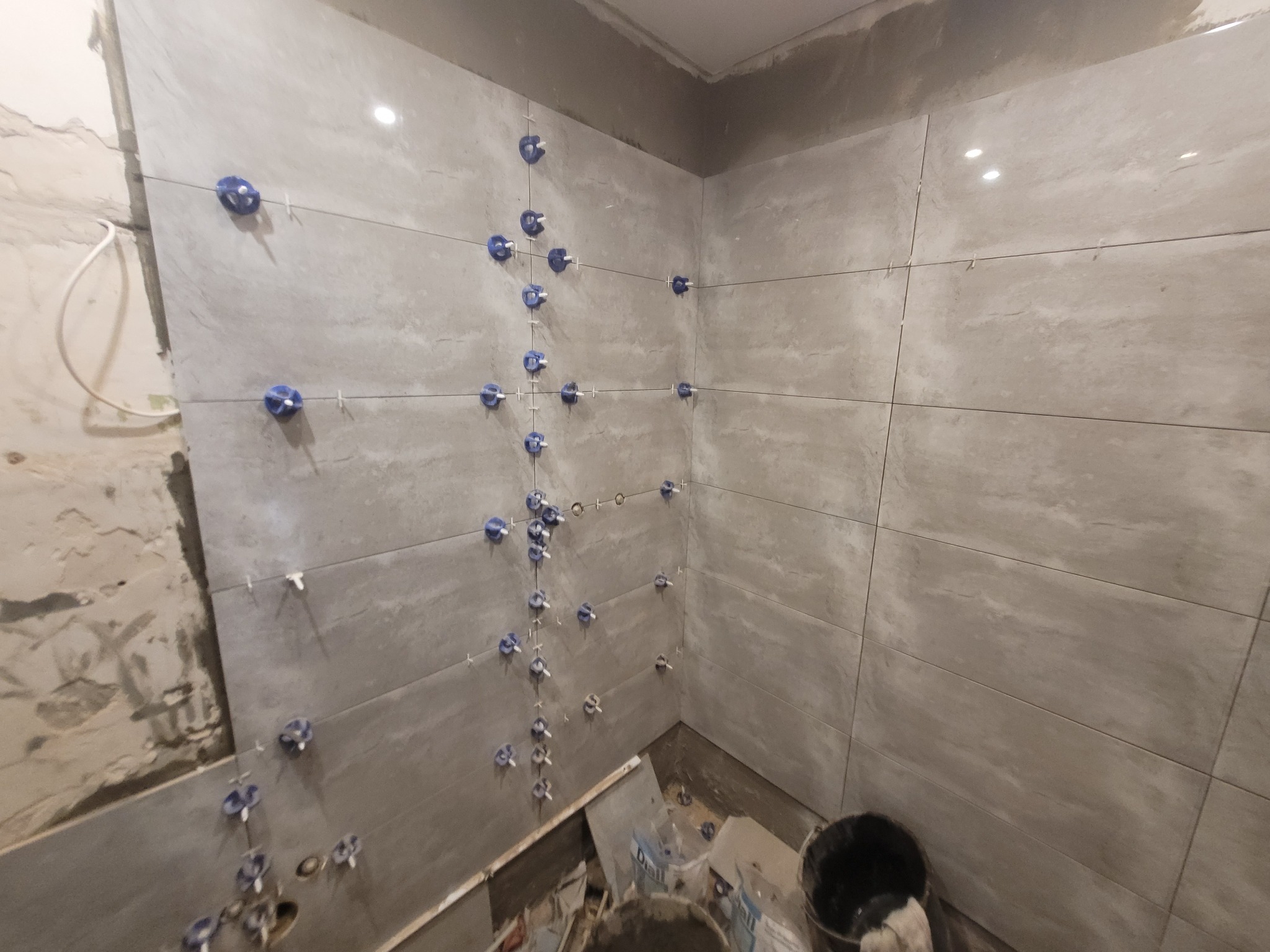 Ściana łazienki w trakcie układania szarych płytek ceramicznych, z widocznymi niebieskimi klipsami poziomującymi i krzyżykami dystansowymi, obok fragment nieotynkowanej ściany i wiadro z zaprawą.