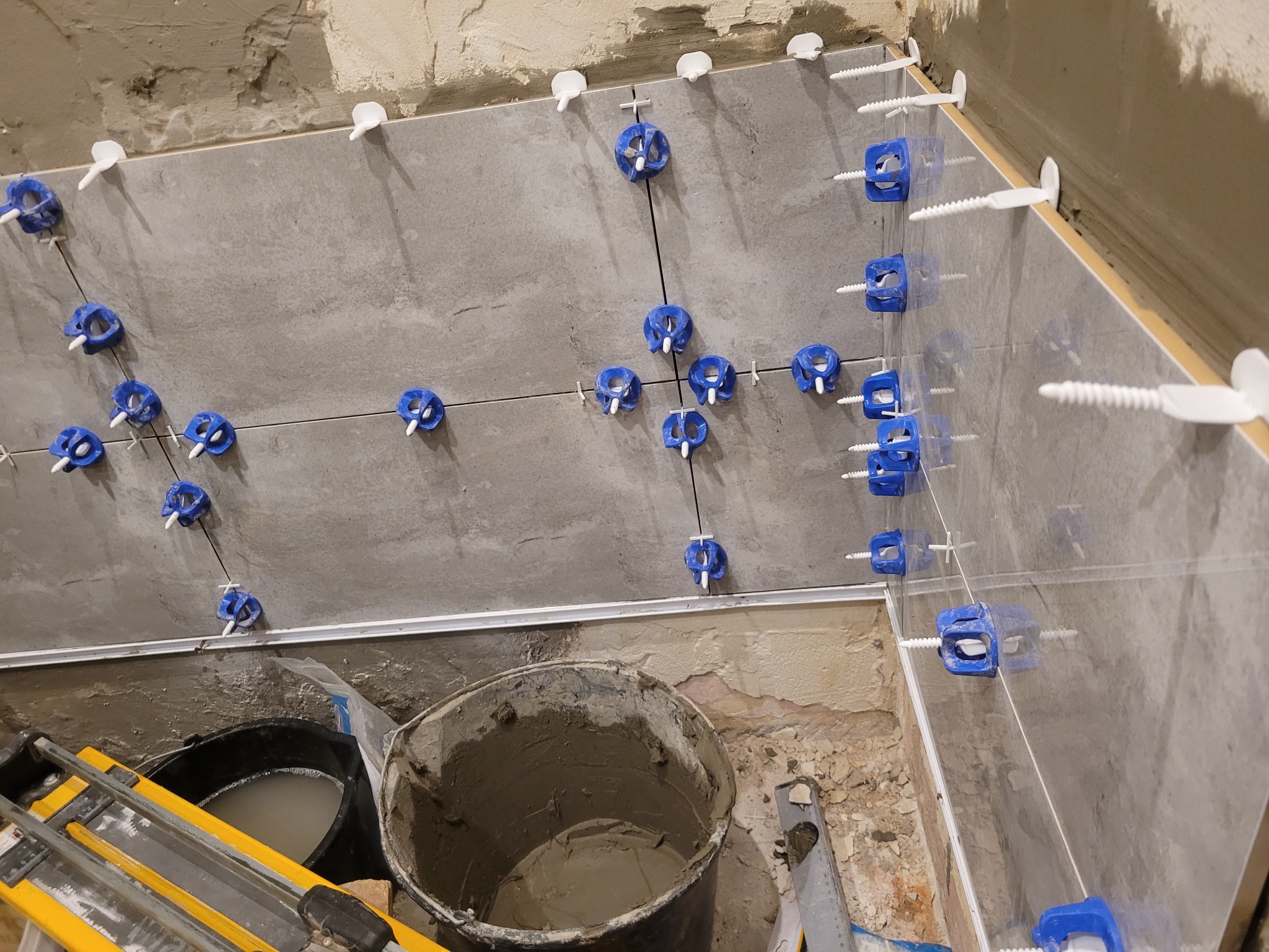 Świeżo ułożone, szare płytki w łazience, wyrównane za pomocą niebieskich klipsów i białych krzyżyków dystansowych. Widoczne surowe ściany i narzędzia glazurnicze w wiadrach.