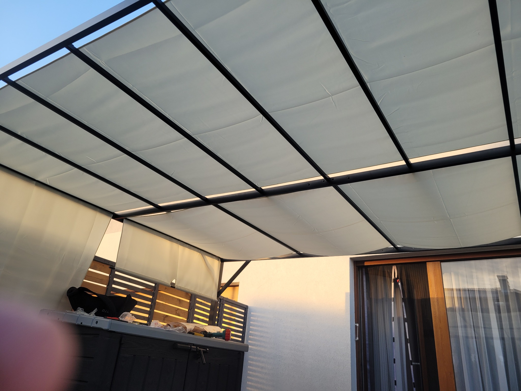 Zadaszenie tarasu z jasną tkaniną i ciemną metalową konstrukcją, widok z dołu na tle ściany budynku i okna przesuwnego.
