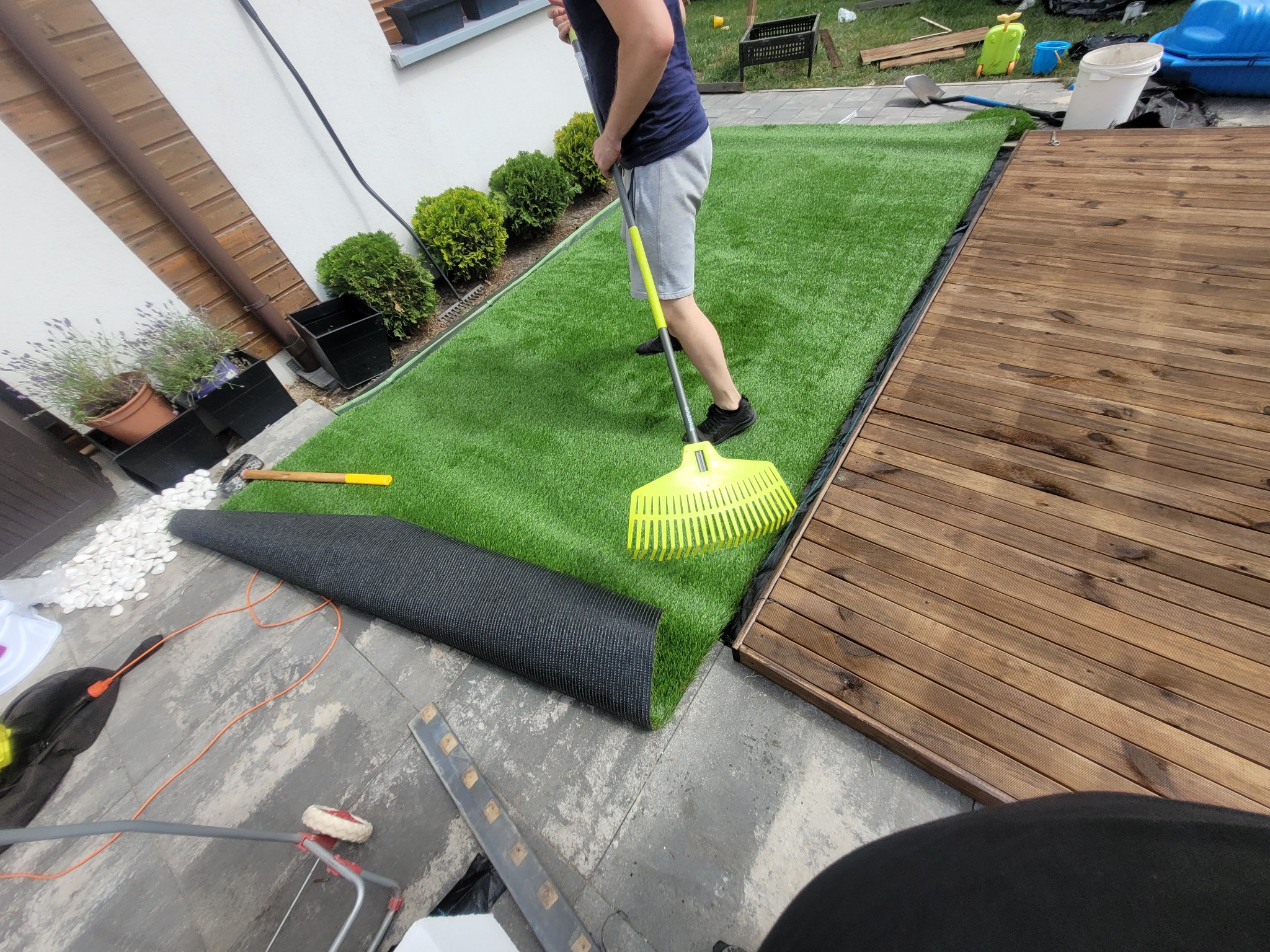 Mężczyzna w szarych spodenkach i granatowej koszulce rozkłada rolkę sztucznej trawy na tarasie z drewnianych desek, wyrównując ją żółtym grabiami. Część trawy leży jeszcze zwinięta na szarym bruku...