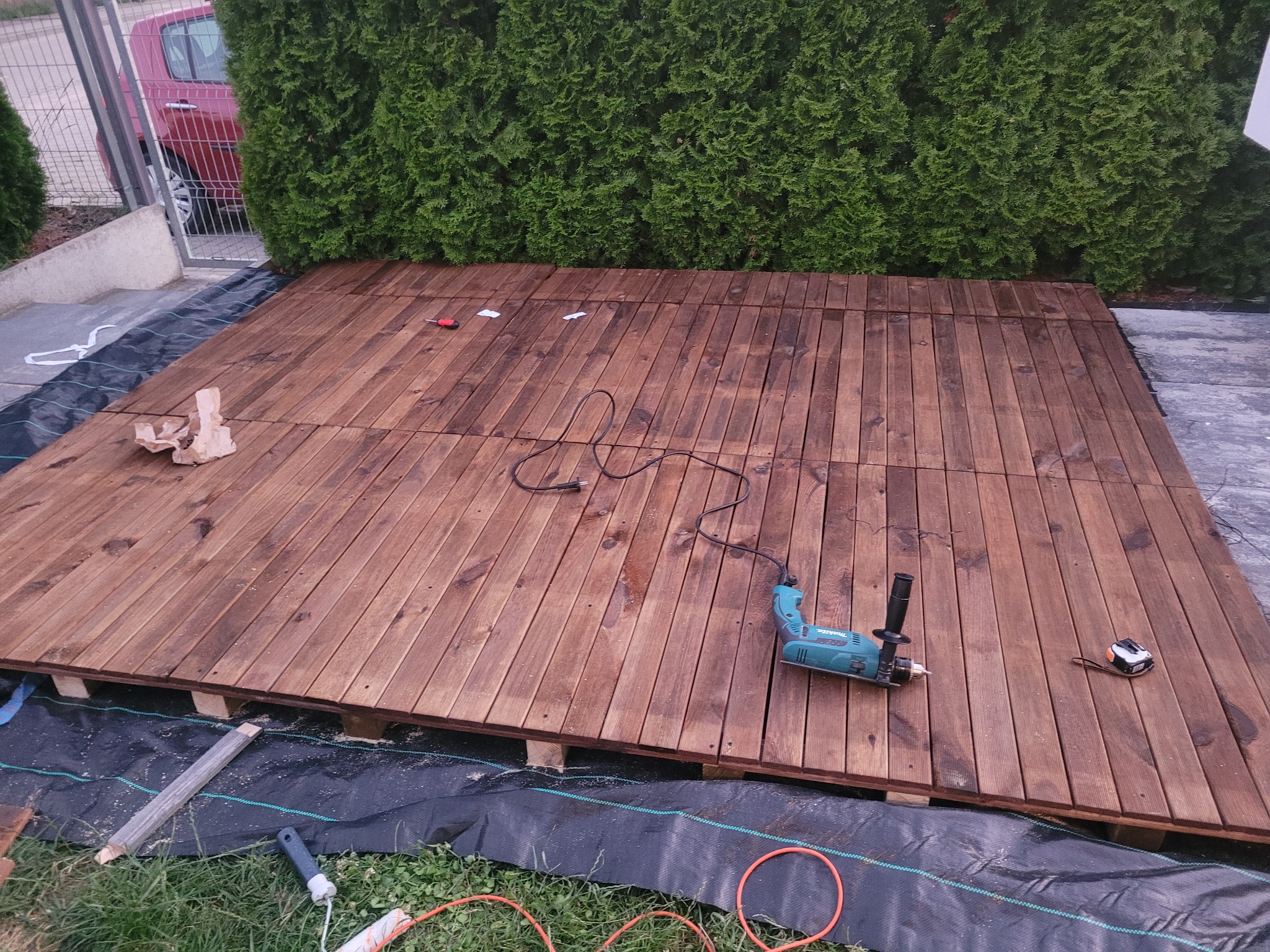 Montaż drewnianego tarasu z desek ryflowanych na czarnej geowłókninie, widoczna wkrętarka akumulatorowa Makita, miara zwijana i częściowo ułożone deski, w tle zielony żywopłot i czerwony samochód.