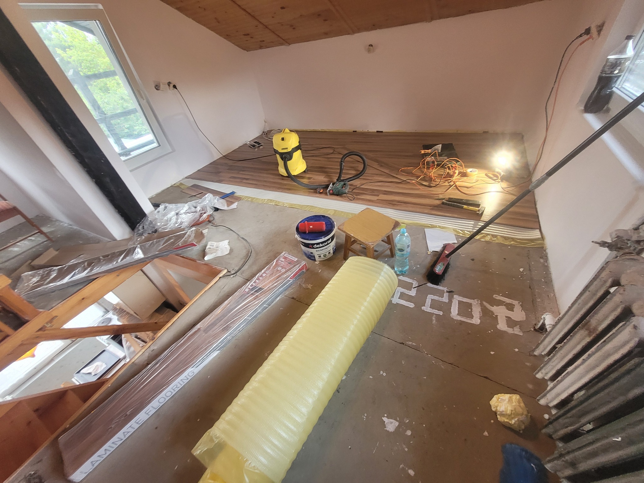 Montaż paneli podłogowych w pokoju na poddaszu. Widoczny odkurzacz przemysłowy, rolka podkładu pod panele, paczki paneli Laminate Flooring, narzędzia, lampa halogenowa oraz fragment grzejnika.