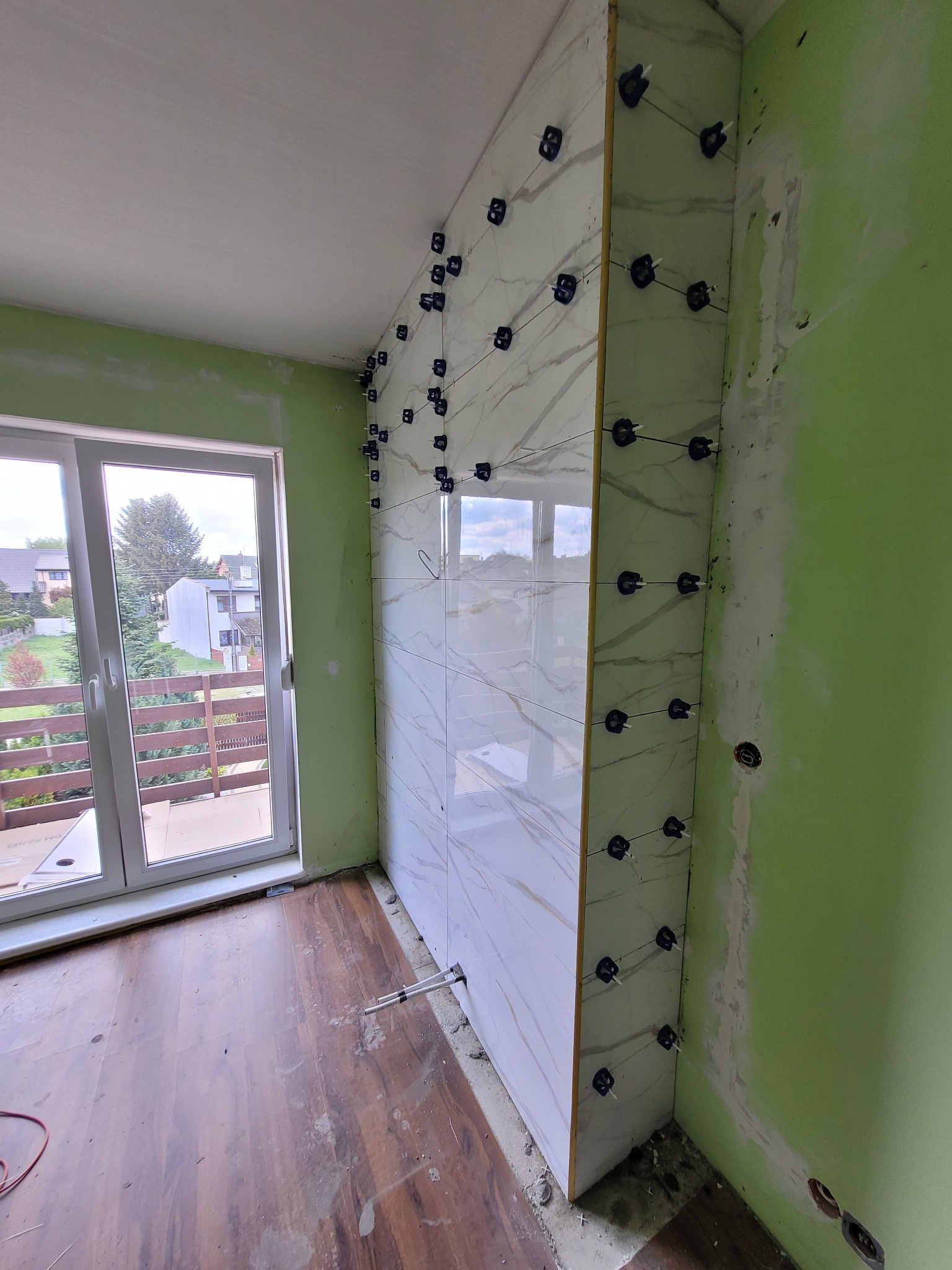 Ściana w trakcie układania płytek imitujących marmur, z użyciem systemu poziomowania, widoczne przewody i niedokończone ściany w pomieszczeniu z oknem balkonowym.