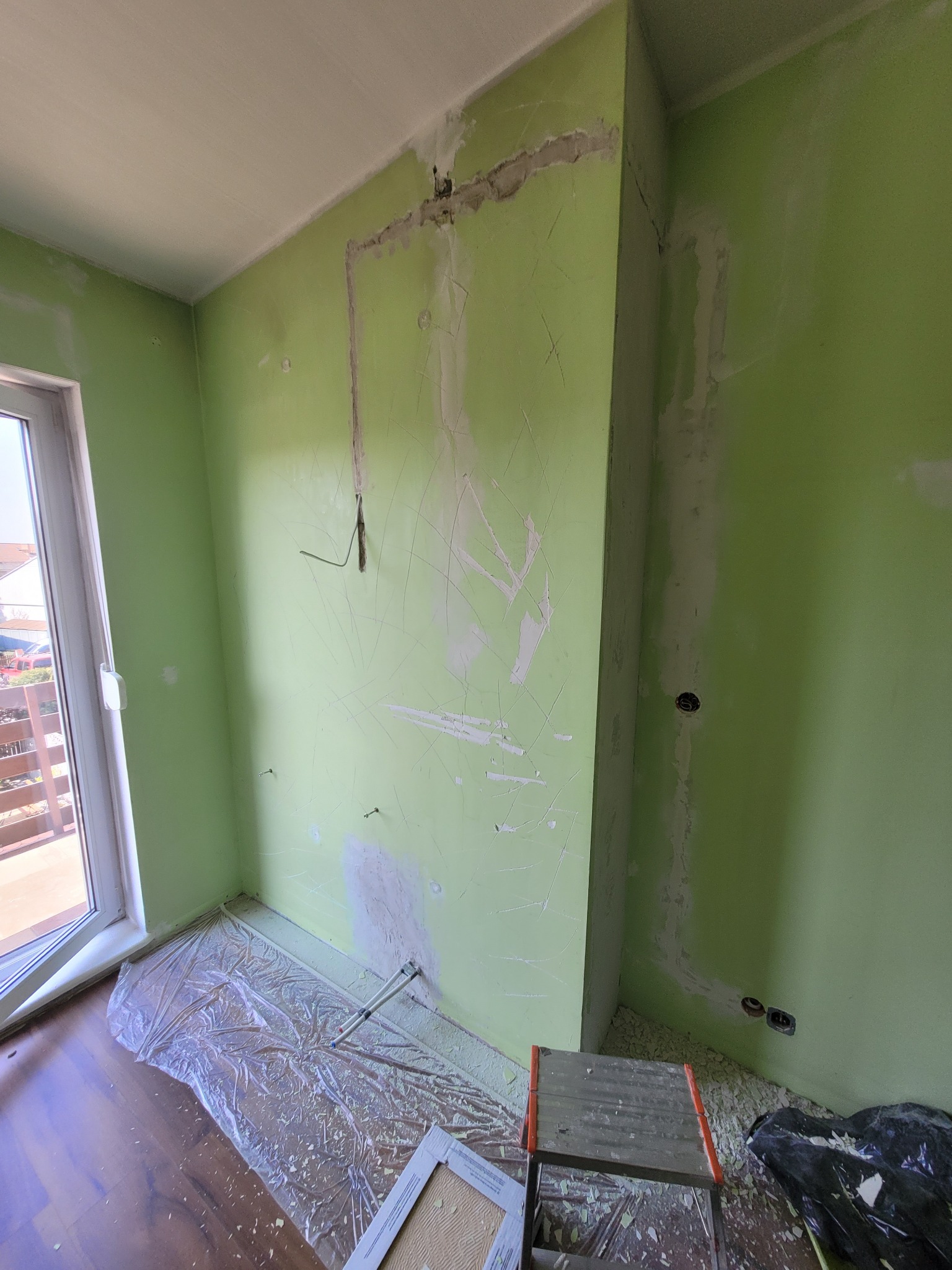 Pokój z zielonymi ścianami w trakcie remontu: widoczne ślady po instalacjach elektrycznych, częściowo zagruntowane ściany, folia ochronna na podłodze i drabina malarska.