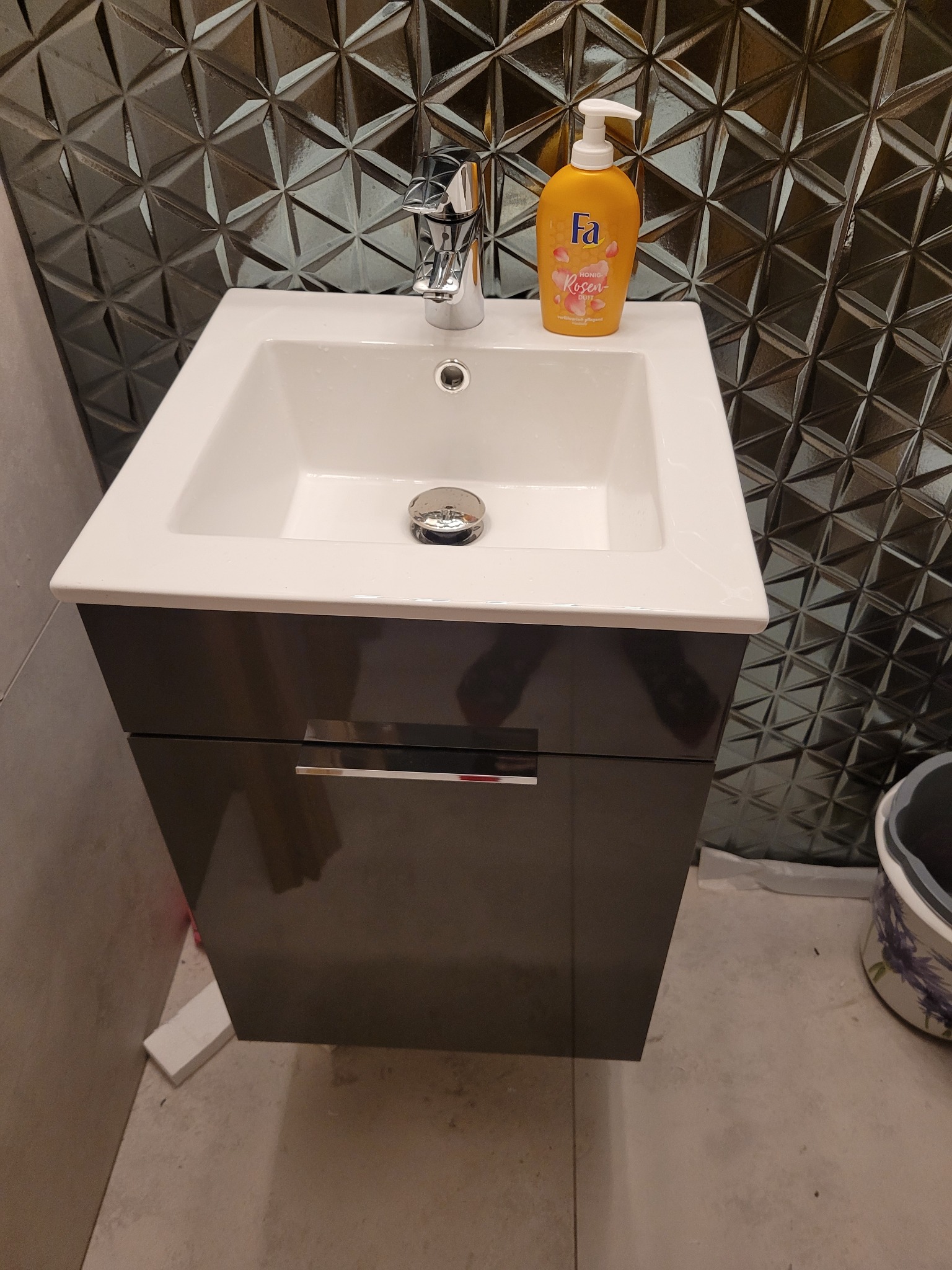 Nowoczesna umywalka nablatowa z szafką w ciemnym kolorze, chromowana bateria i dozownik mydła Fa Rosen na tle ściany z trójwymiarowych płytek w geometryczny wzór.