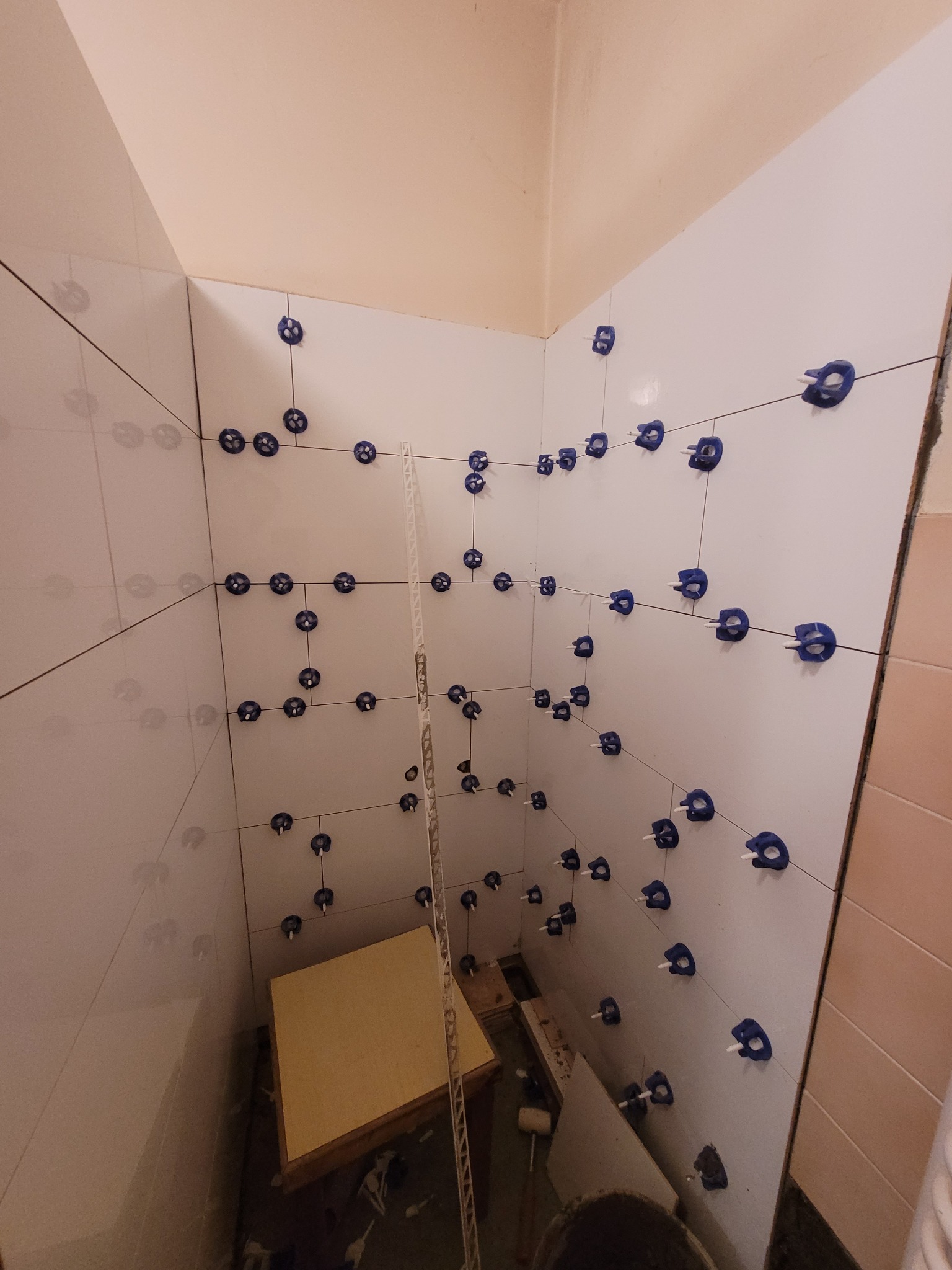 Świeżo ułożone, białe płytki w rogu łazienki, z użyciem niebieskich klipsów i klinów poziomujących, widoczna aluminiowa listwa narożna i fragment wiadra na podłodze.