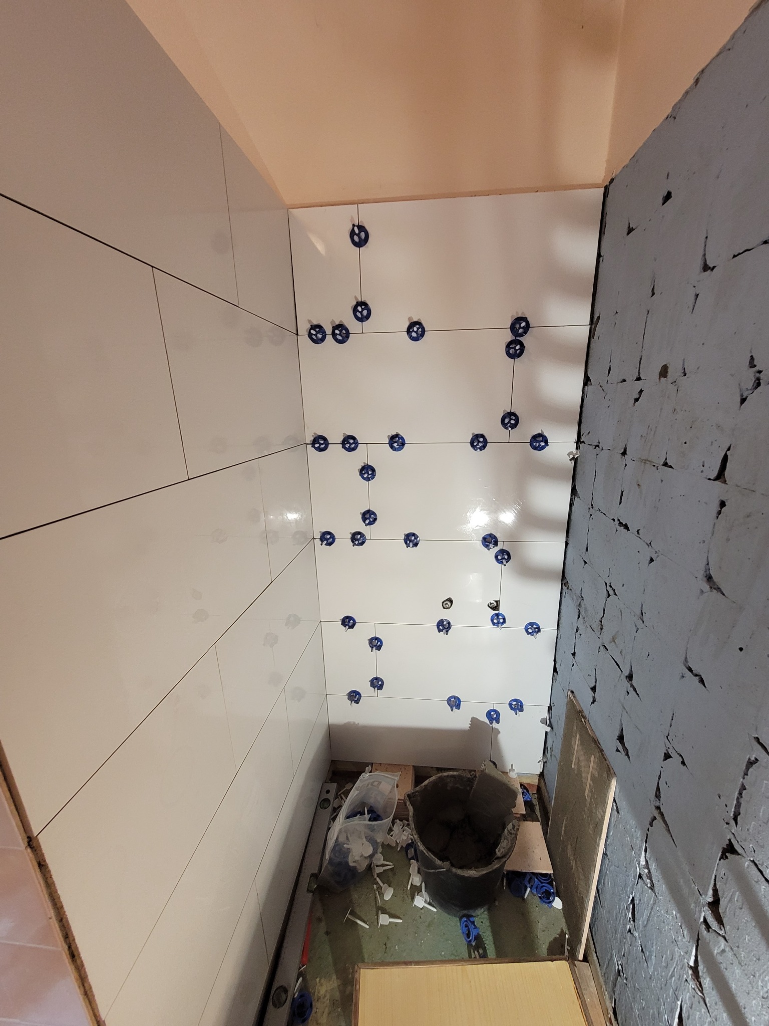 Świeżo ułożone białe płytki ścienne w łazience, zabezpieczone niebieskimi krzyżykami dystansowymi, sąsiadują z surową, szarą ścianą przygotowaną pod dalsze prace wykończeniowe; widoczne wiadro...