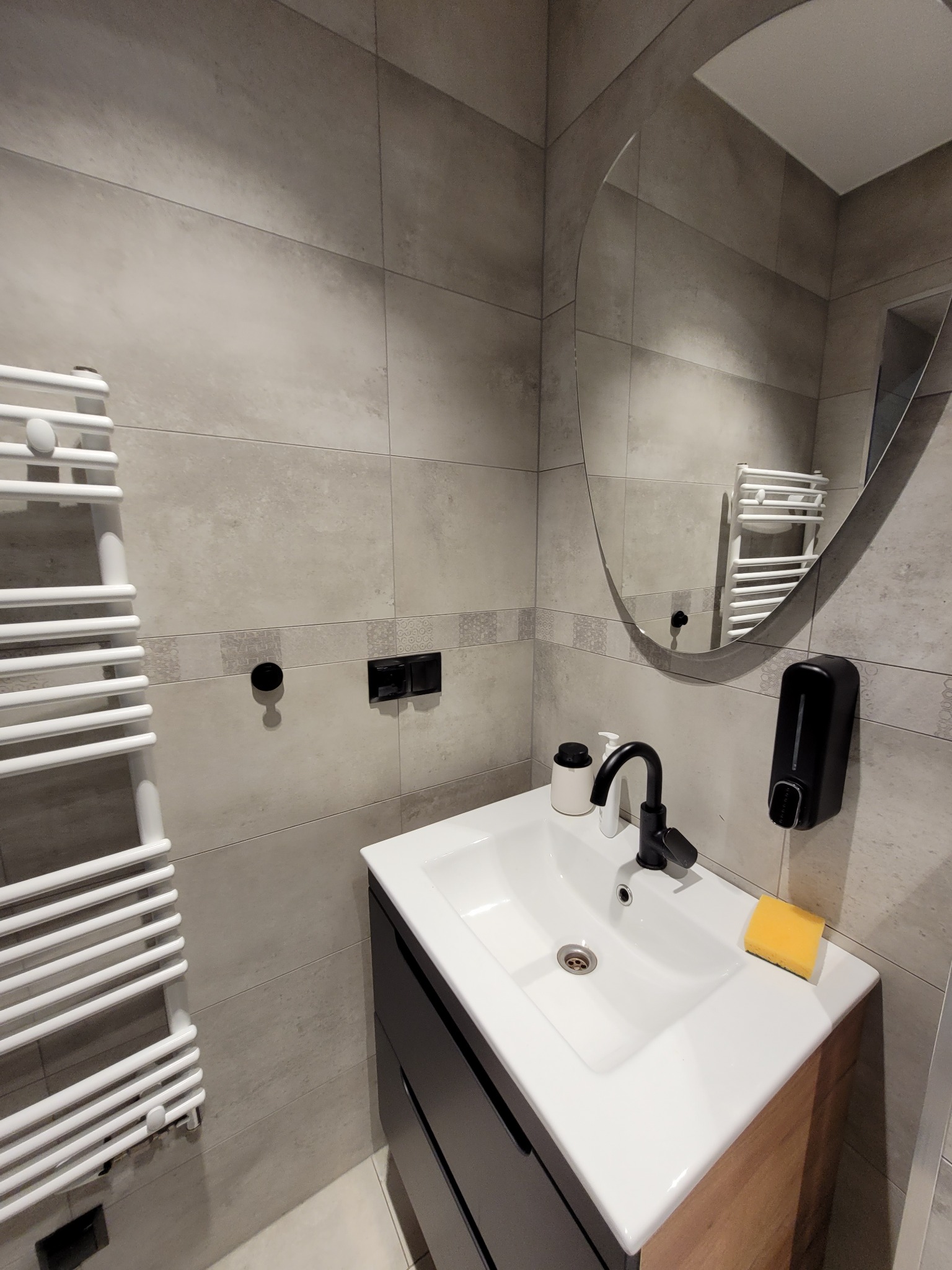 Nowoczesna łazienka z szarymi płytkami, owalnym lustrem, białą umywalką z czarną armaturą, grzejnikiem drabinkowym i dozownikiem mydła w czarnym kolorze.