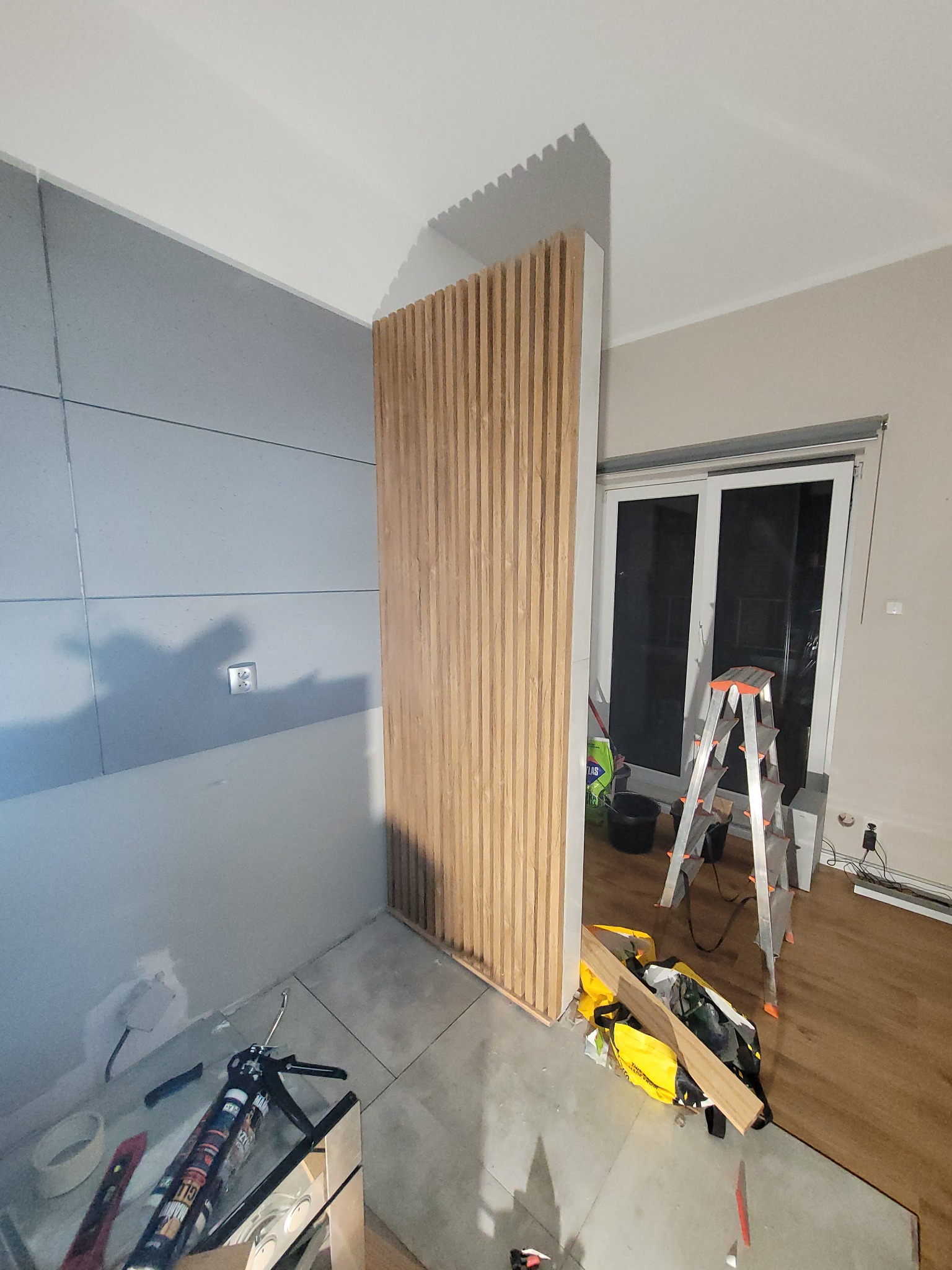 Montaż drewnianej ścianki działowej z lameli w pomieszczeniu z przygotowaną ścianą z płyt gipsowych, widoczne narzędzia i drabina.
