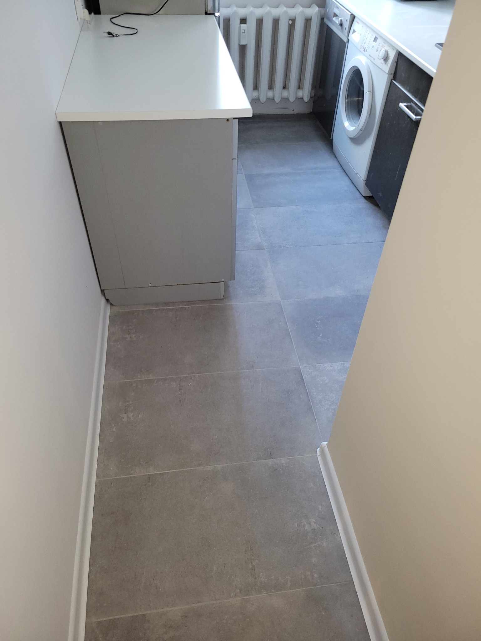 Szare płytki podłogowe ułożone w łazience z widoczną pralką, grzejnikiem i fragmentem szarej szafki pod umywalkę z białym blatem.