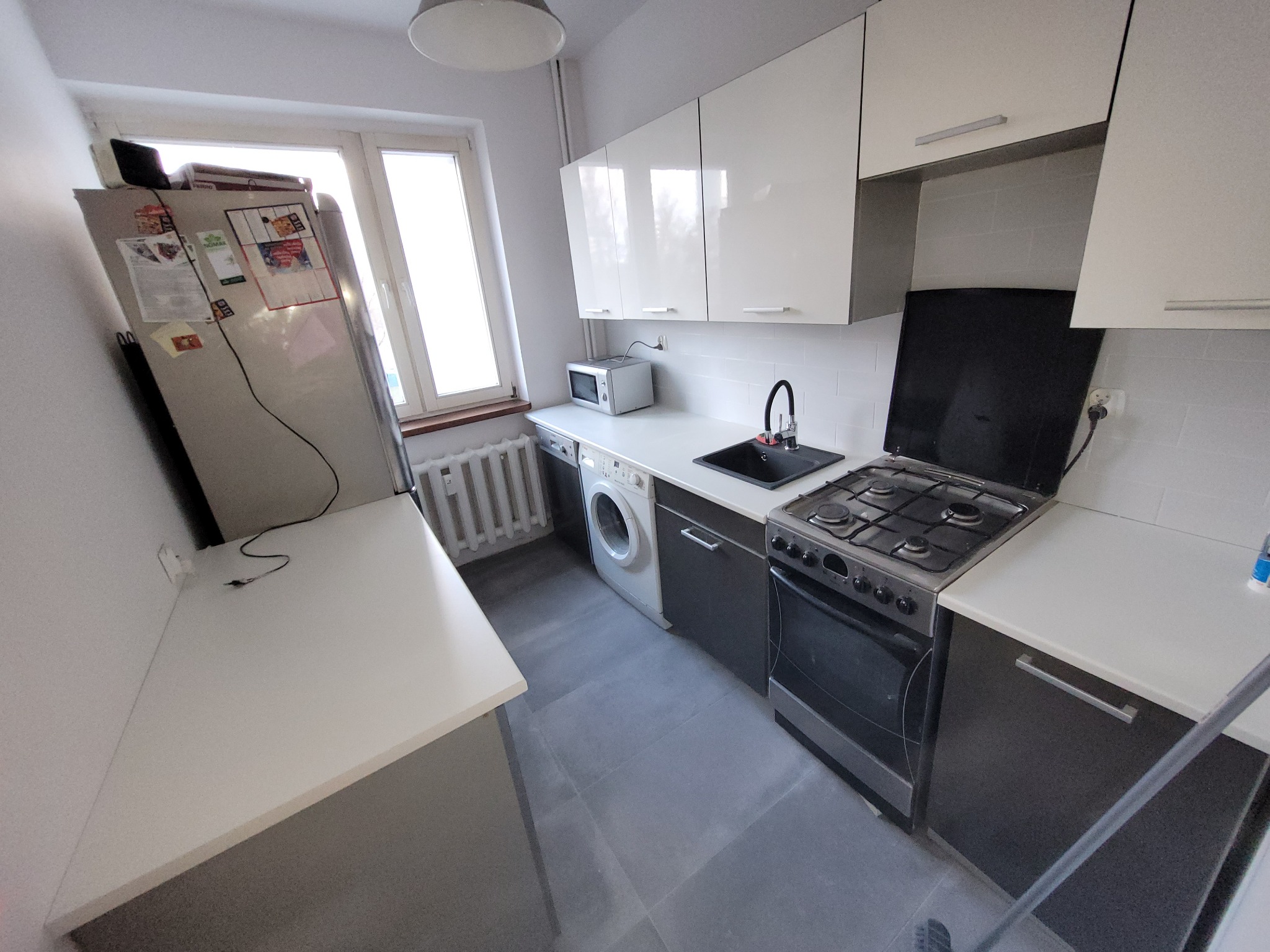 Wnętrze małej kuchni w bloku z białymi szafkami, szarą podłogą i sprzętami AGD; widoczna pralka pod blatem, kuchenka gazowa i lodówka z magnesami na drzwiach.