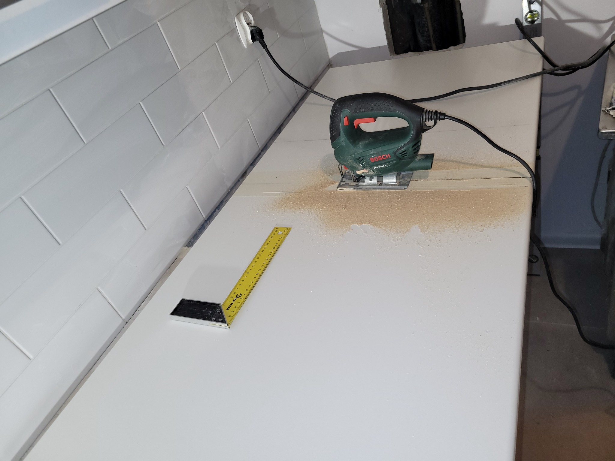 Zielony wyrzynarka Bosch PST 7200 E tnie biały blat w łazience wyłożonej białymi płytkami, obok leży żółty kątownik TOP TOOLS, widoczne ślady cięcia i pył.