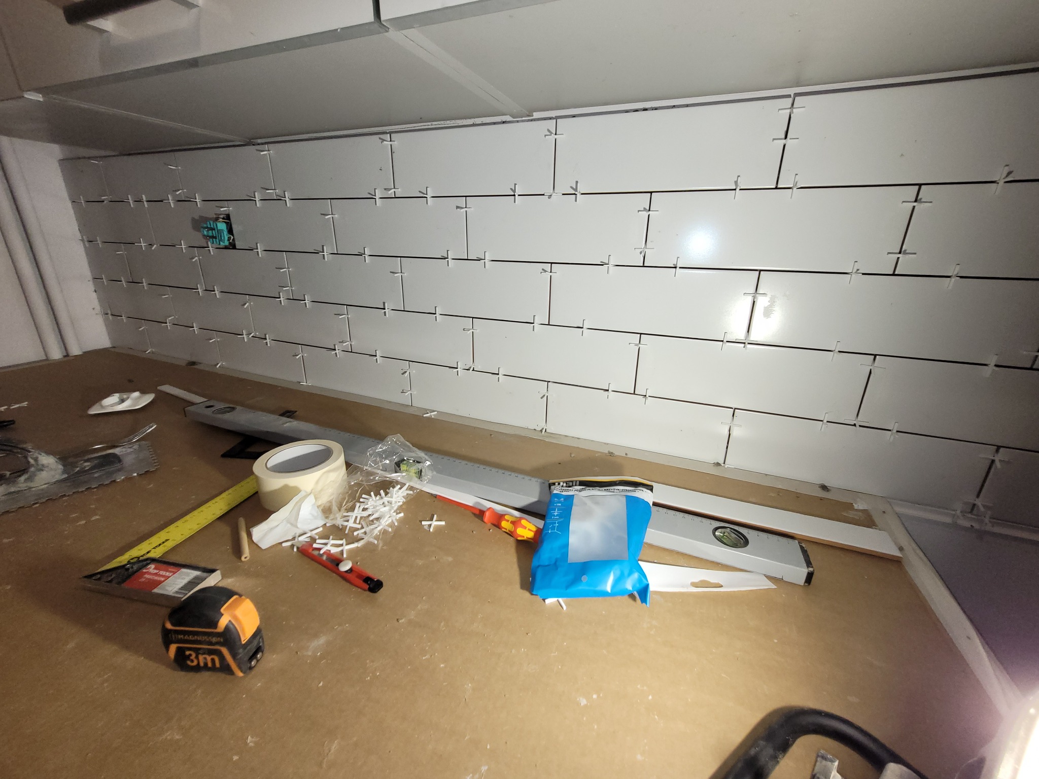 Ściana z białych płytek, w trakcie układania, z krzyżykami dystansowymi, widoczne narzędzia: poziomica, miarka, taśma, szpachelka i opakowanie z krzyżykami.
