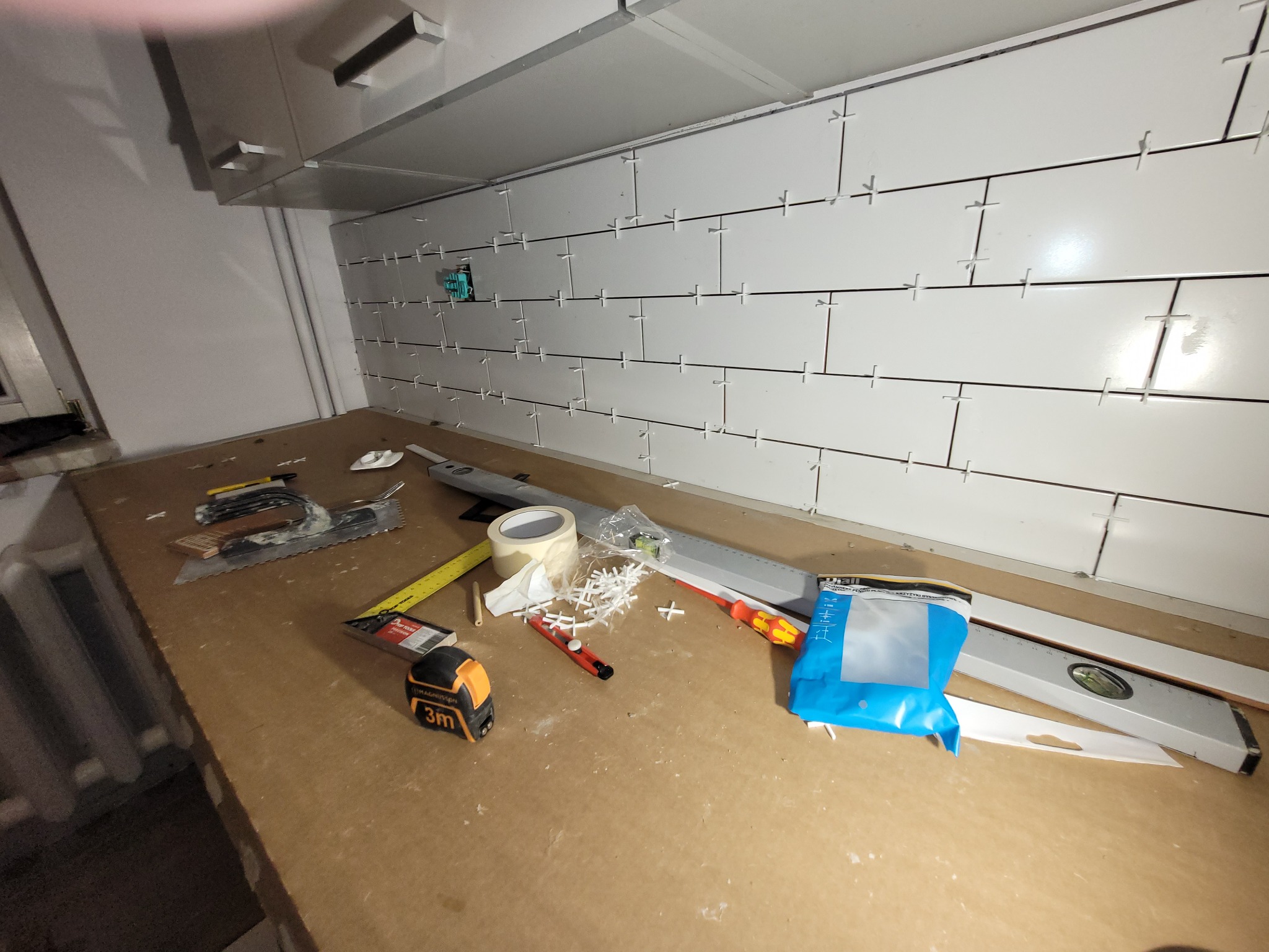 Ściana z białych płytek ułożonych w cegiełkę, z plastikowymi krzyżykami dystansowymi między nimi, widoczne narzędzia glazurnicze: poziomica, miara, nóż, paca zębata, szpachelka i taśma malarska.