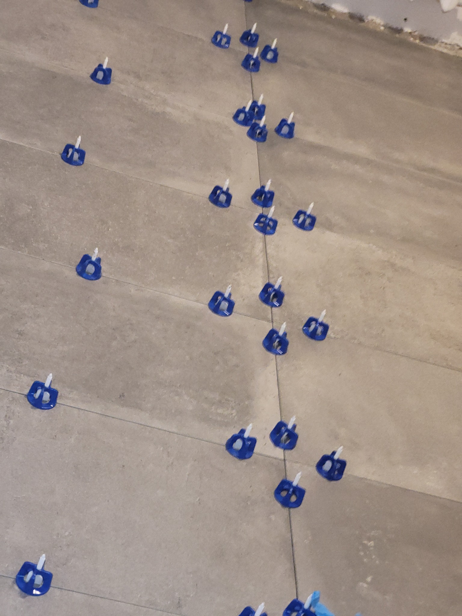 Ułożone płytki podłogowe w trakcie montażu, z użyciem niebieskich klipsów poziomujących i sznurka, widoczne niedociągnięcia na ścianie w tle.