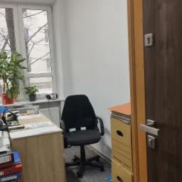 Biuro w Lublinie: Kobieta pracuje przy biurku z komputerem, obok okno z widokiem na drzewa, za nią regał z segregatorami.