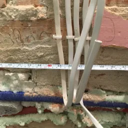 Przewody elektryczne i rury wodne zabezpieczone pianką montażową na tle ceglanej ściany z widocznymi śladami po pracach instalacyjnych i miarą zwijaną.
