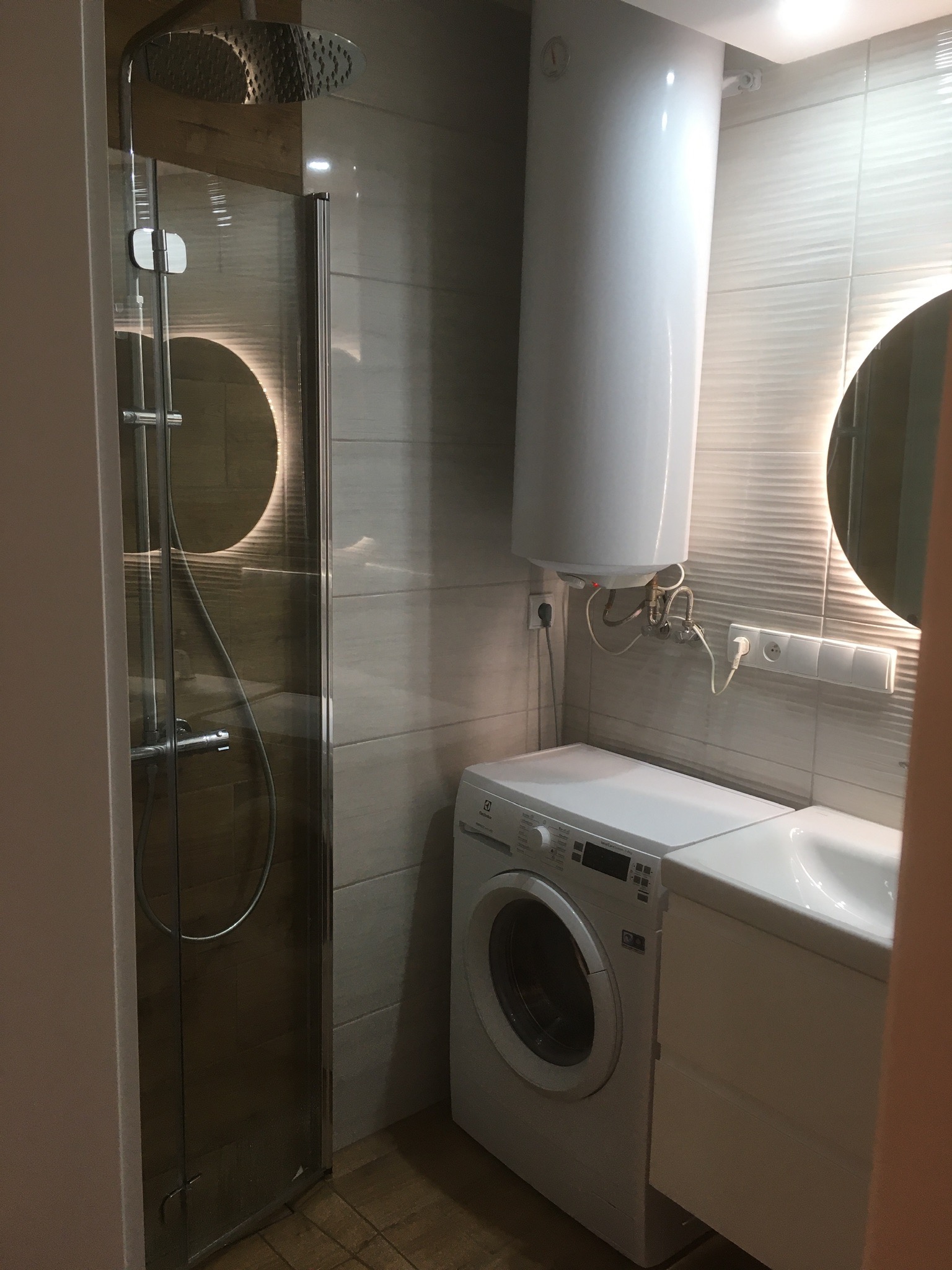 Nowoczesna łazienka z kabiną prysznicową, okrągłym lustrem z podświetleniem LED, pralką Electrolux i bojlerem elektrycznym.