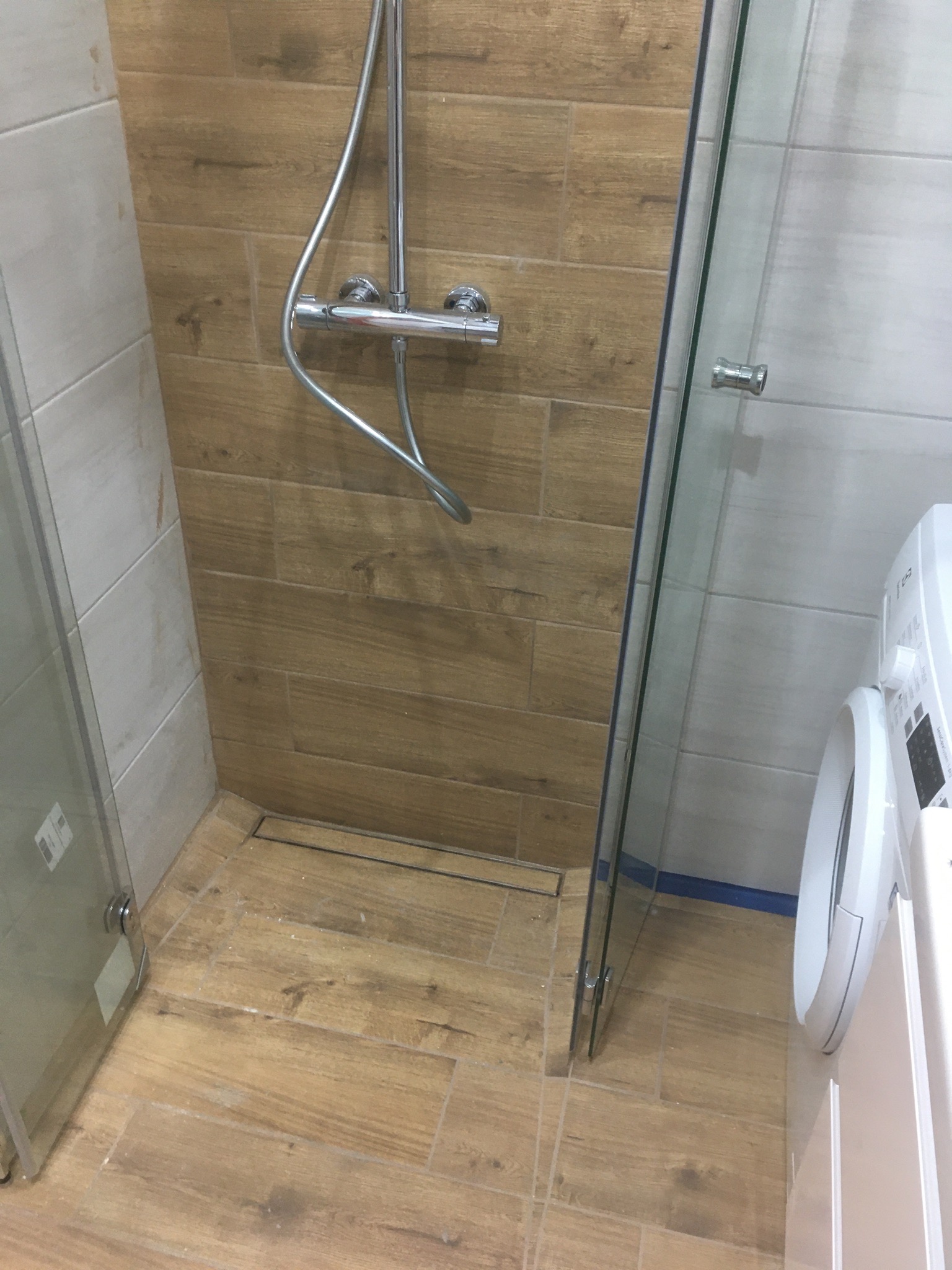 Kabina prysznicowa z odpływem liniowym, wykończona płytkami imitującymi drewno, obok pralka