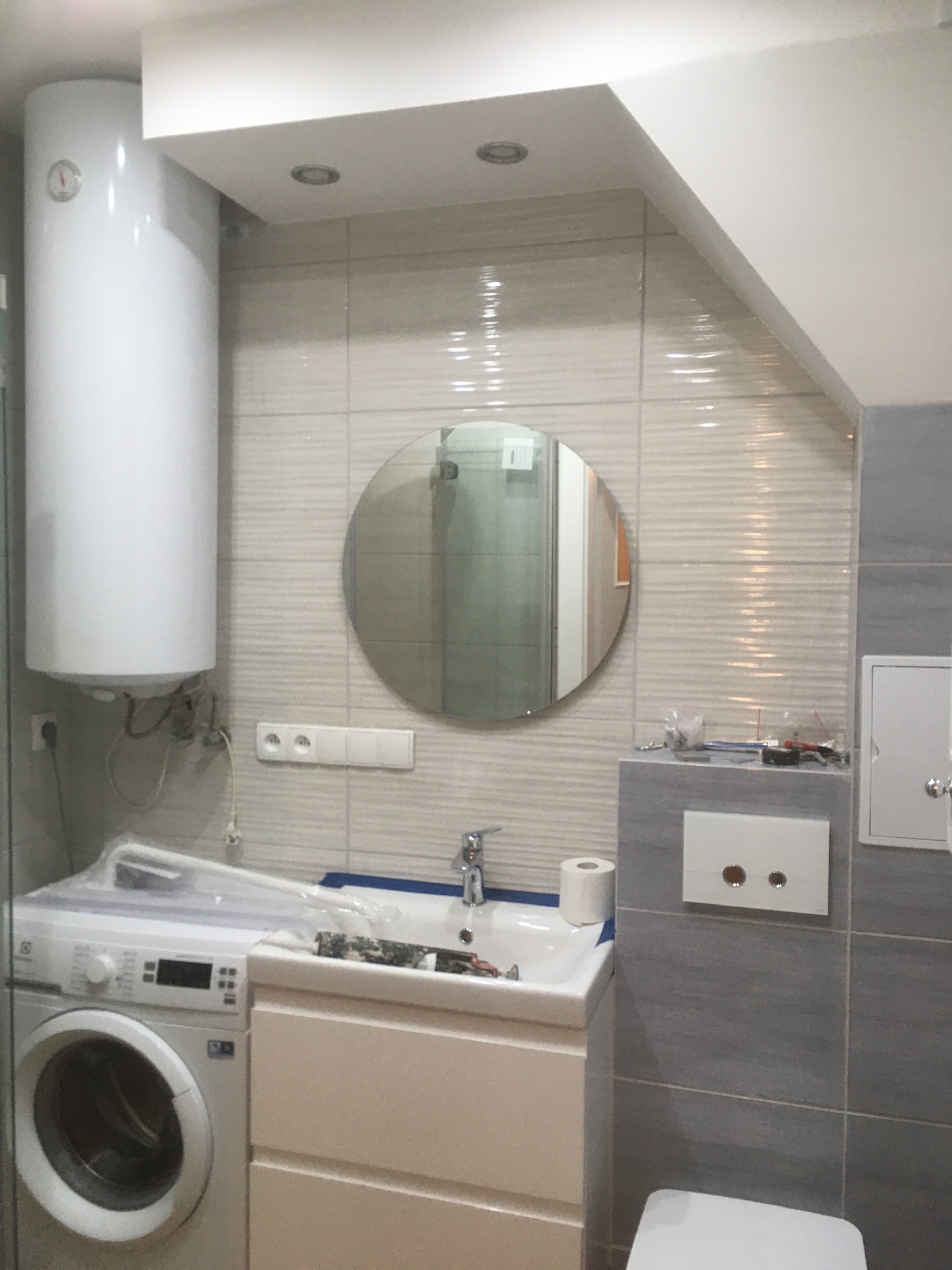 Łazienka w trakcie prac wykończeniowych: widoczny bojler, pralka, umywalka z armaturą, okrągłe lustro, płytki w odcieniach szarości, narzędzia na blacie, oraz częściowo widoczna toaleta.
