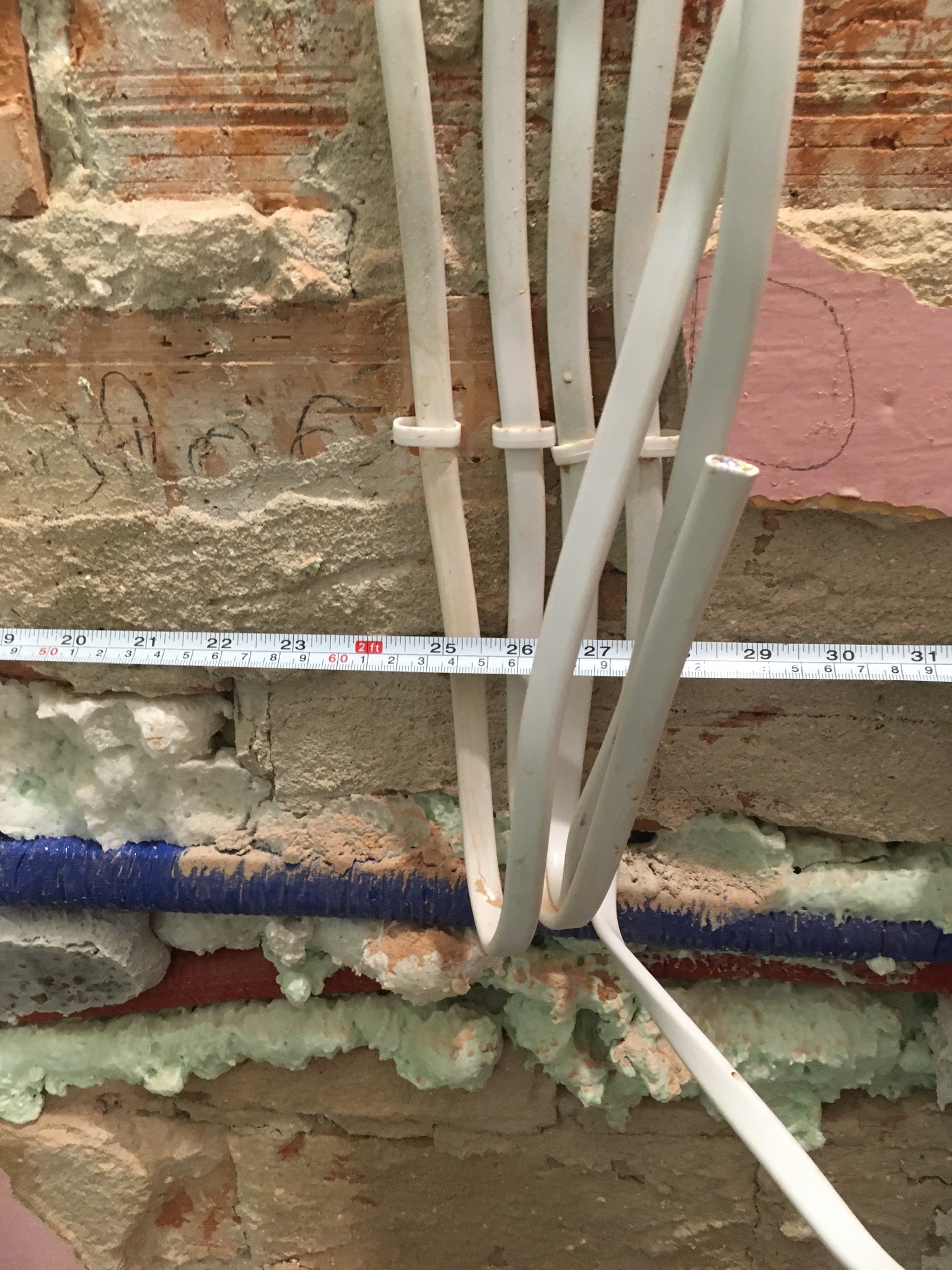 Przewody elektryczne i rury wodne zabezpieczone pianką montażową na tle ceglanej ściany z widocznymi śladami po pracach instalacyjnych i miarą zwijaną.
