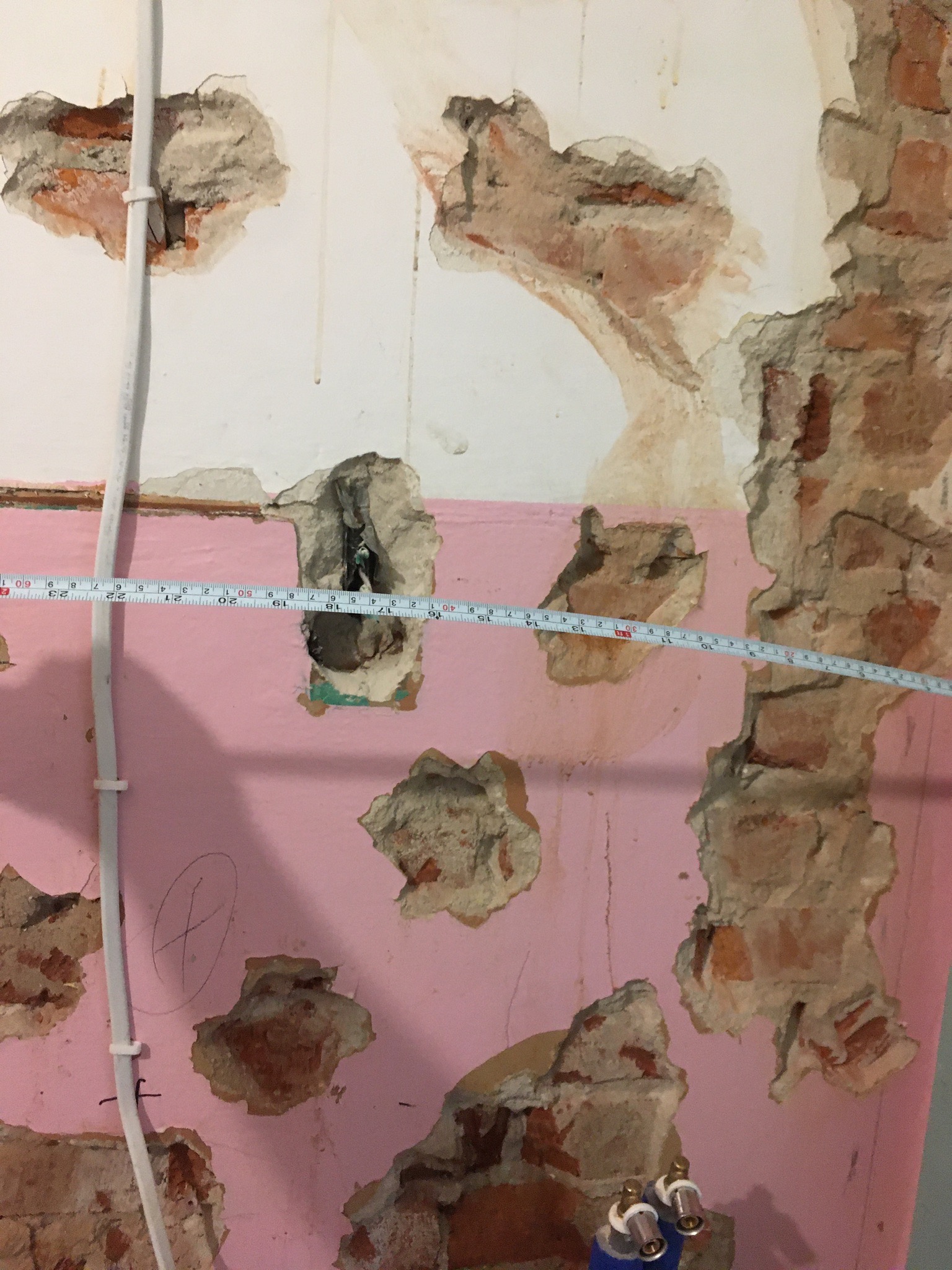 Różowa ściana z uszkodzeniami, odsłoniętą cegłą i rurami wodnymi; pomiar odległości taśmą mierniczą.