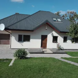 Dom jednorodzinny z szarą blachodachówką, widoczna brama garażowa, drzwi wejściowe z wybrukowanym podejściem i zadbany trawnik.