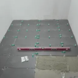 Układanie ciemnych płytek podłogowych z użyciem poziomicy, krzyżyków dystansowych i klipsów poziomujących. Na podłożu rozprowadzona zaprawa klejowa.